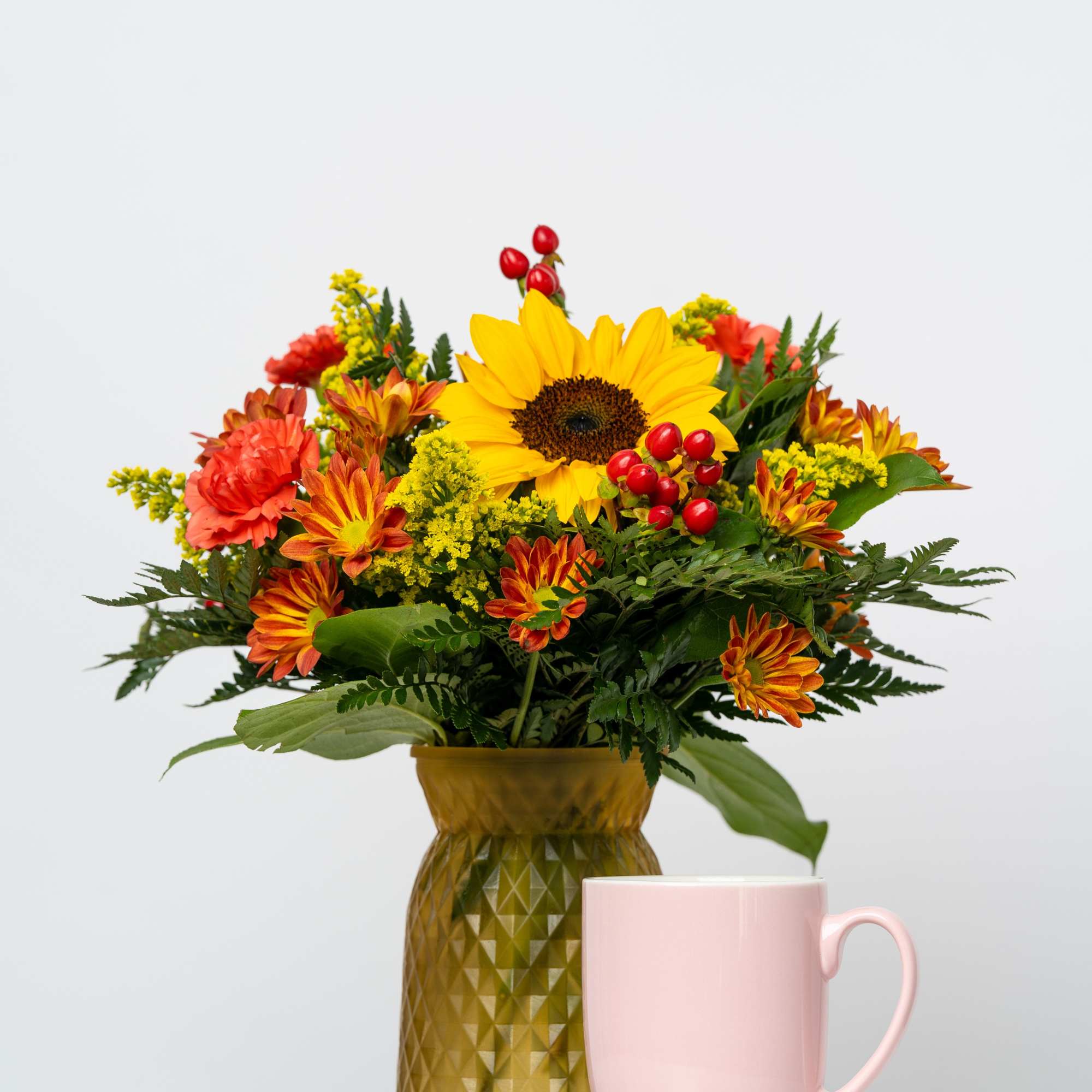 Sunflower and autumn flower arrangement in a gold vase beside a pink mug