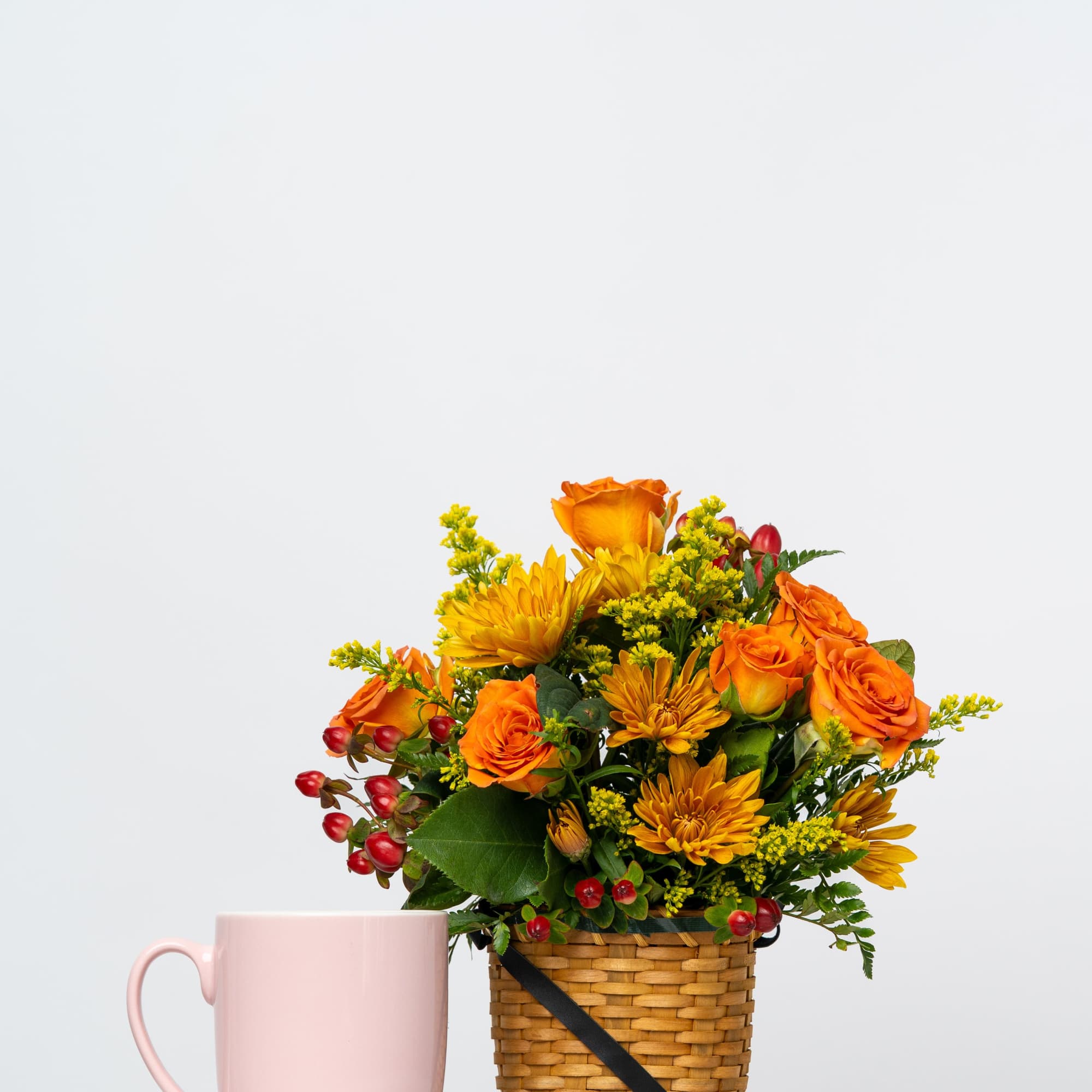 Orange and yellow flower arrangement in a woven basket beside a pink mug