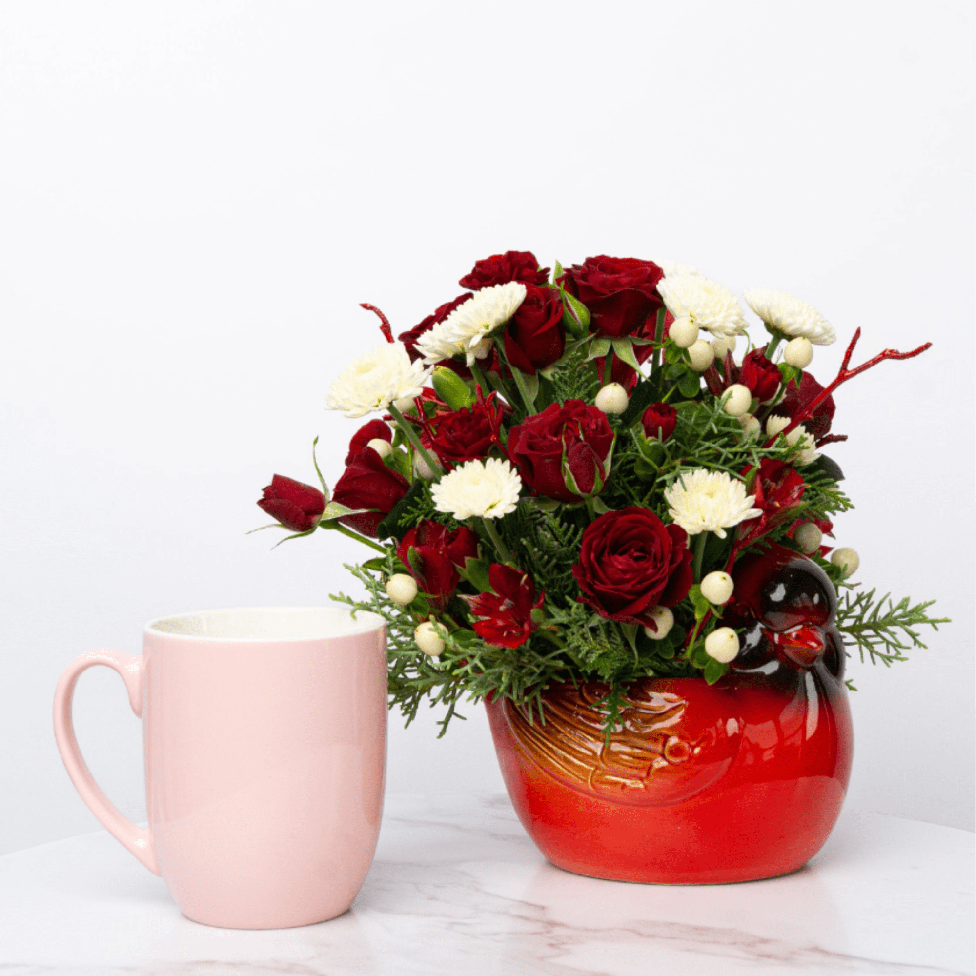 Red and white flower arrangement in a red ceramic mug beside a pink mug