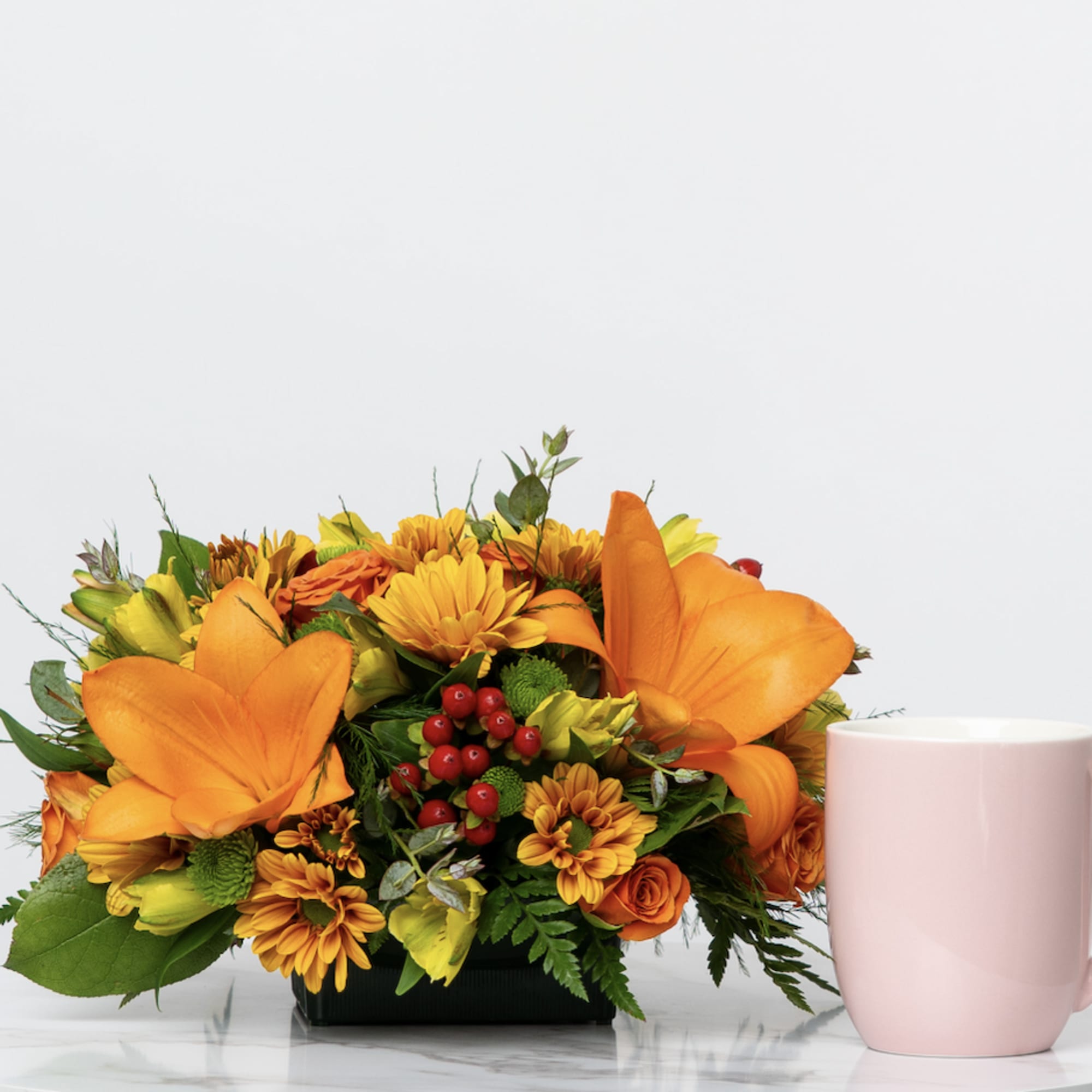 Orange floral arrangement in a black container beside a pink mug