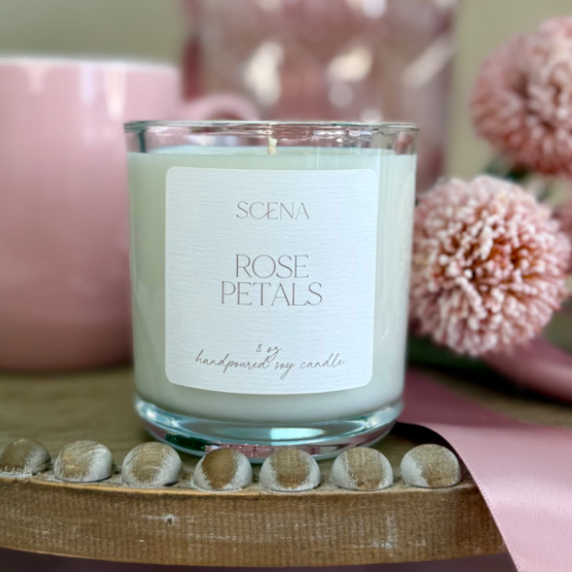 Rose-scented white candle in a clear glass jar with pink mug, flowers, and ribbon on a wooden tray