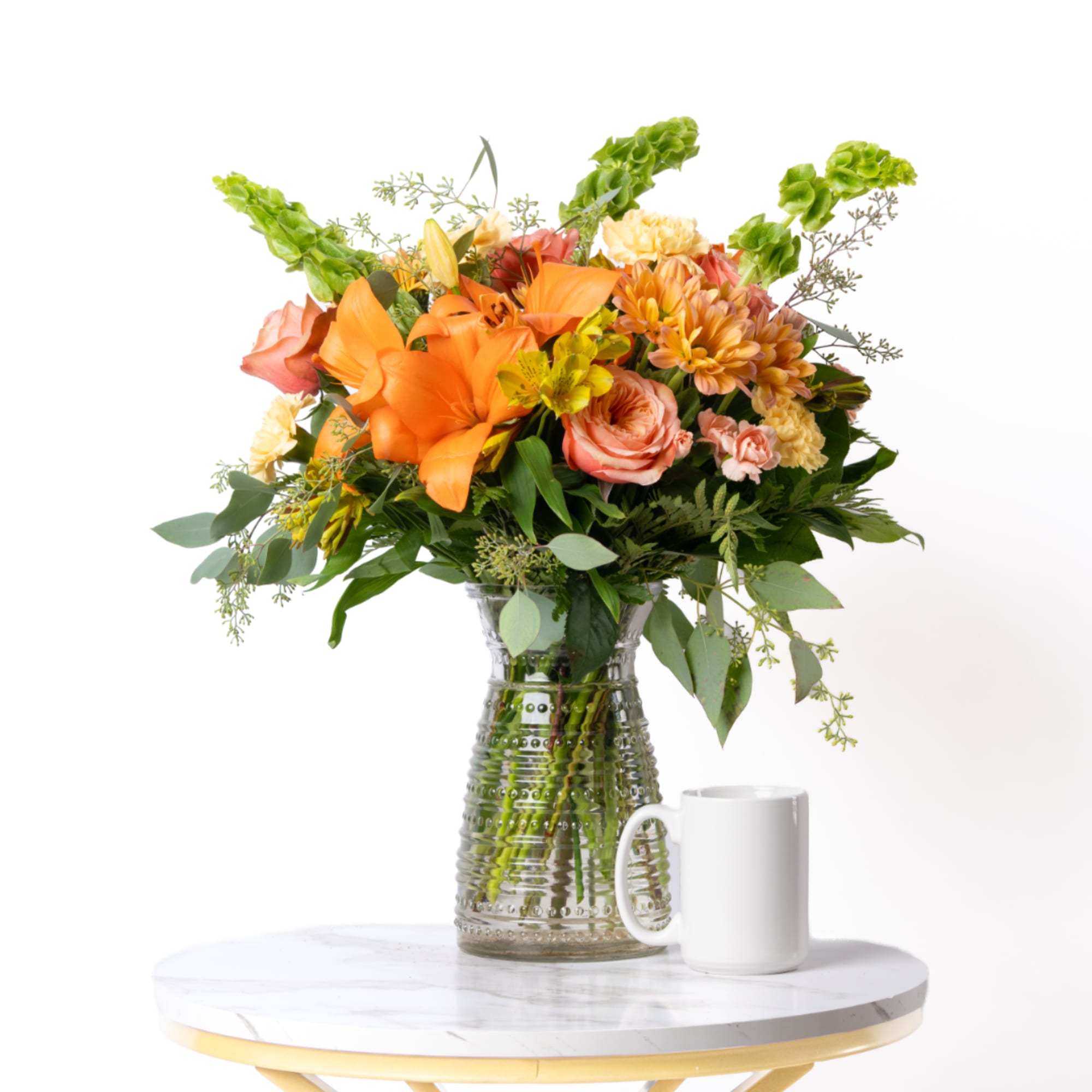 Orange and peach mixed bouquet in a clear glass vase with a white mug beside it.