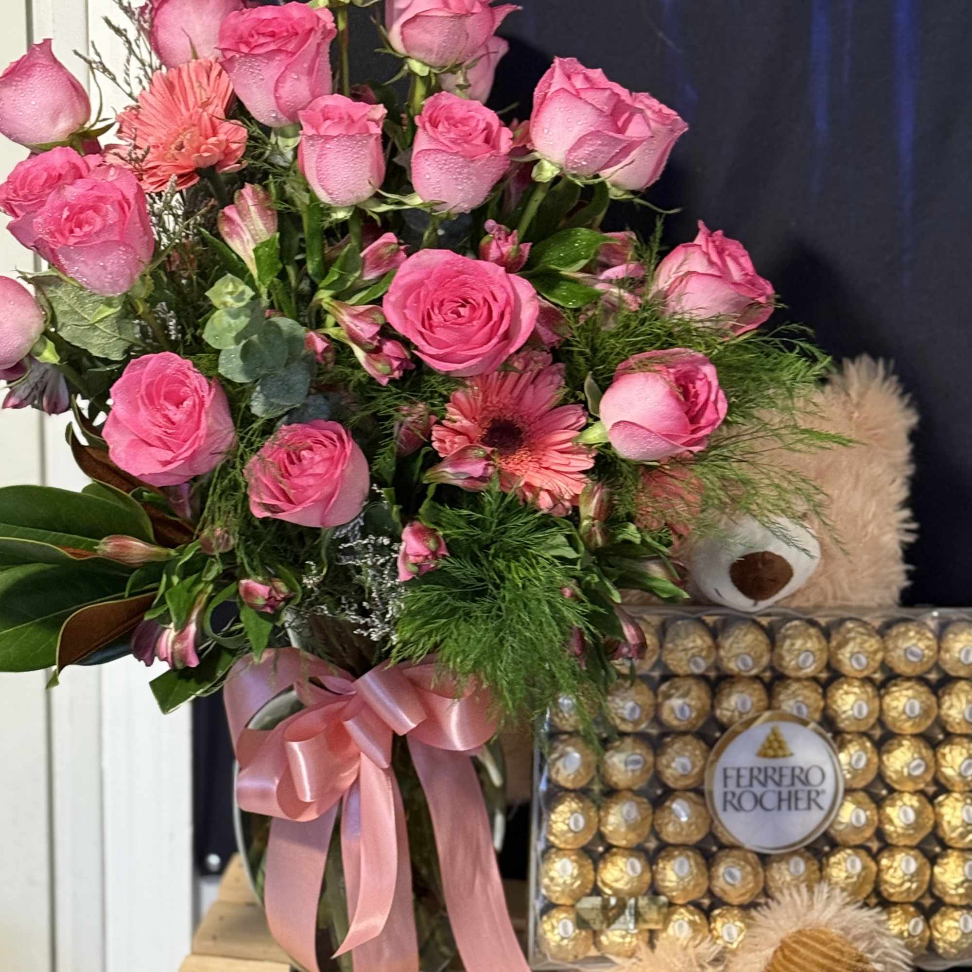Pink rose bouquet in a glass vase beside a Ferrero Rocher box and teddy bear