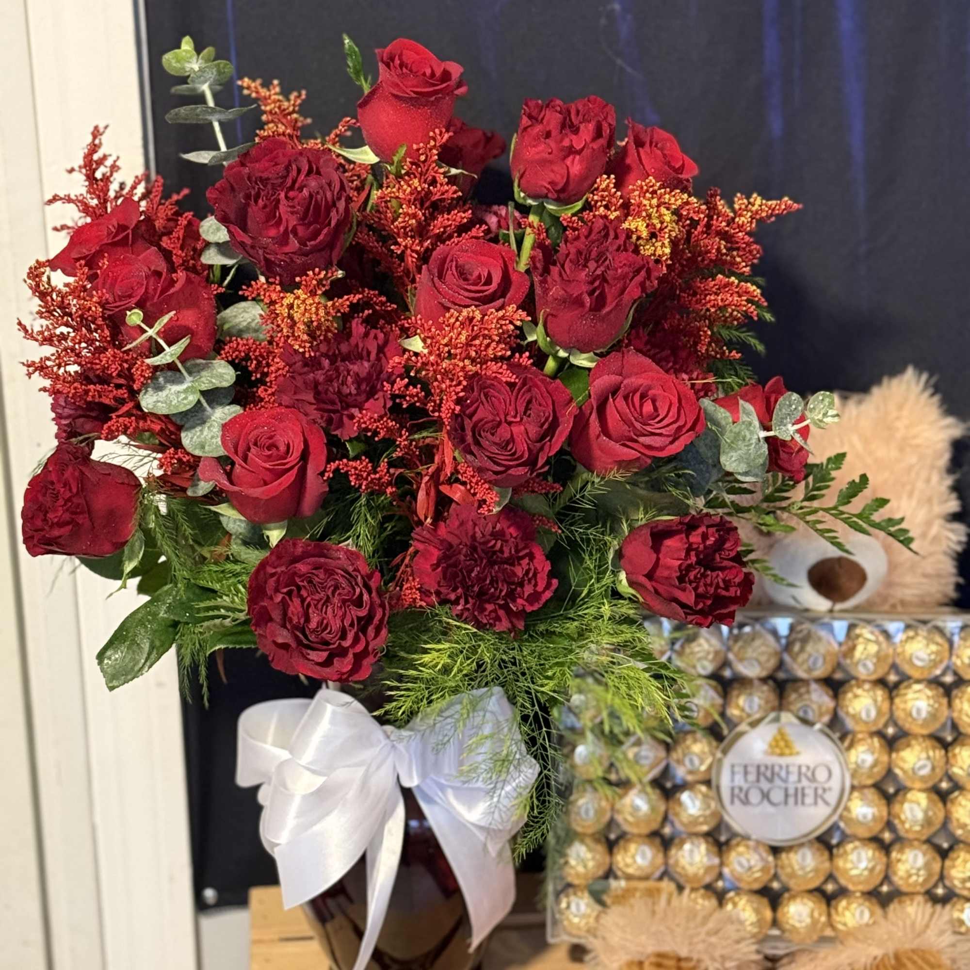 Red rose bouquet in a dark vase with a white ribbon, beside a Ferrero Rocher box and teddy bear