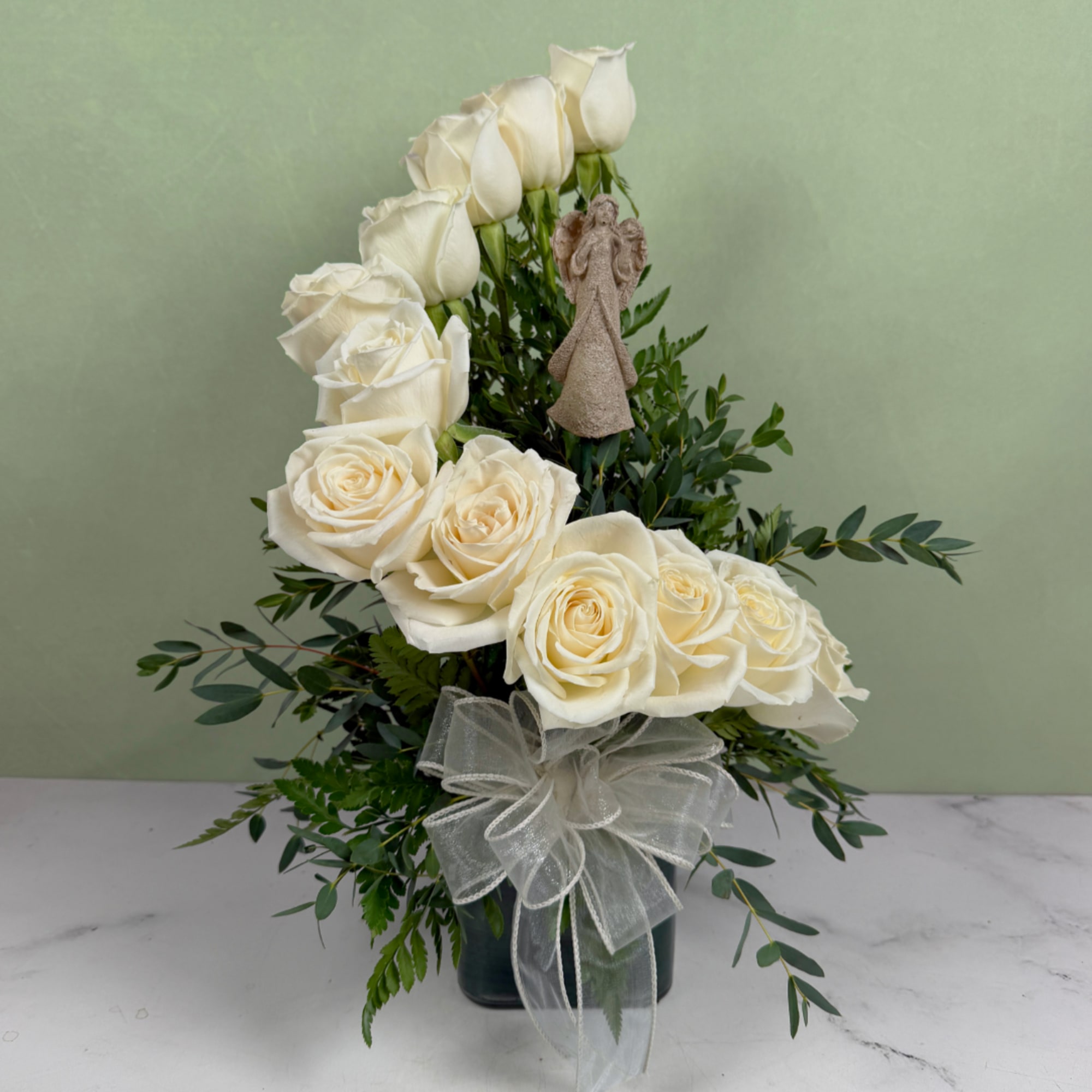 White roses arranged in a vase with greenery, a ribbon, and a small angel figurine