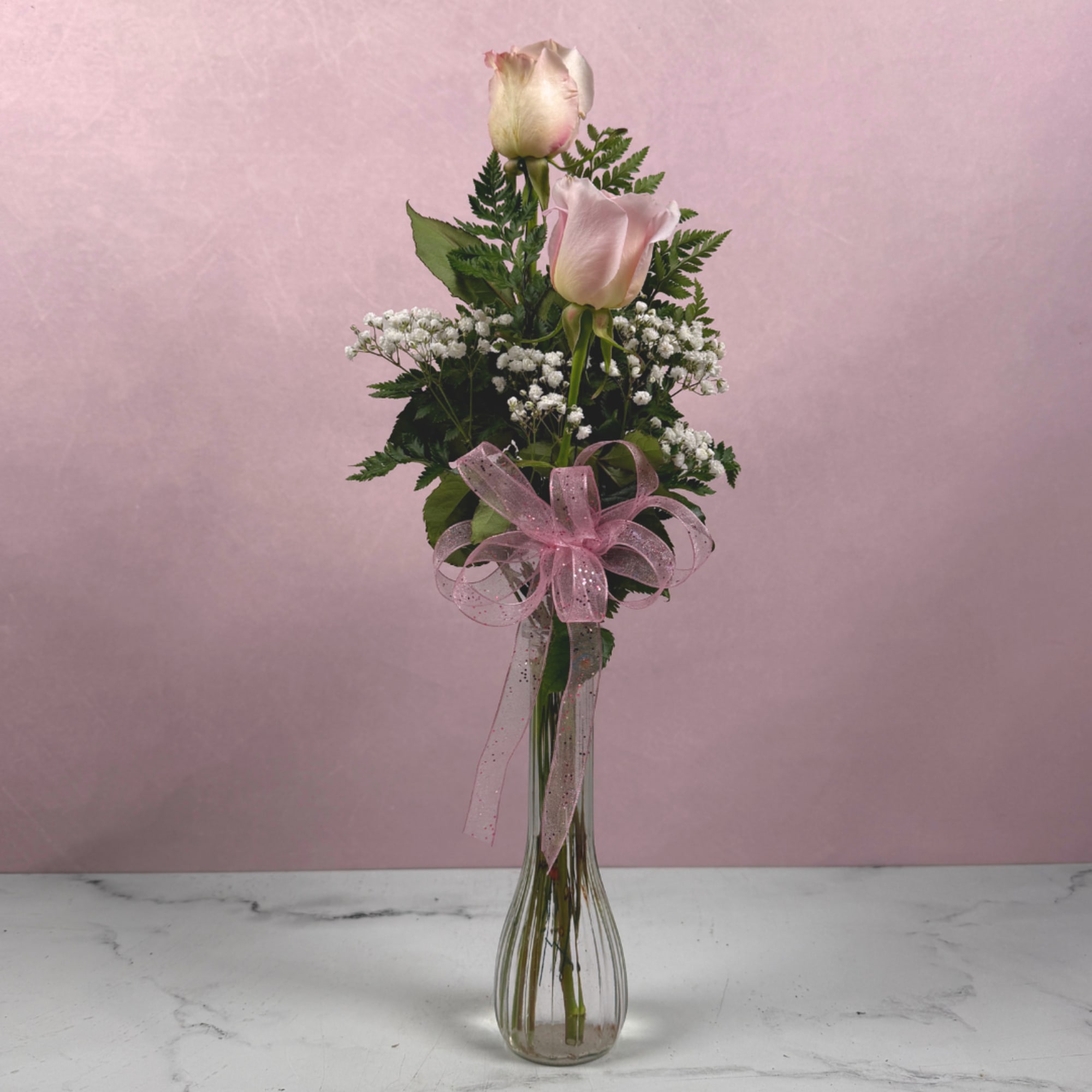 Two pale pink roses in a tall glass vase with baby's breath and a pink ribbon