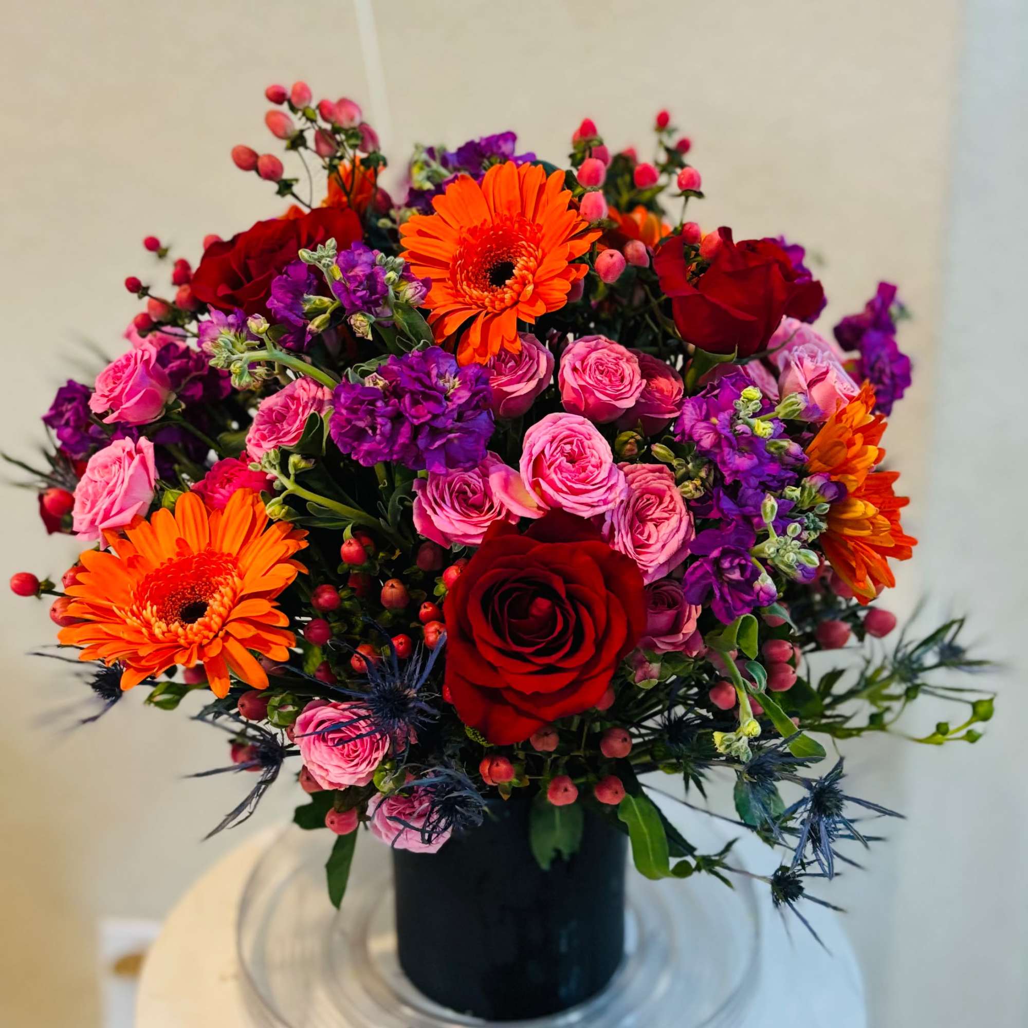 Bright mixed bouquet with red roses, pink spray roses, orange gerberas, and purple blooms in a black vase