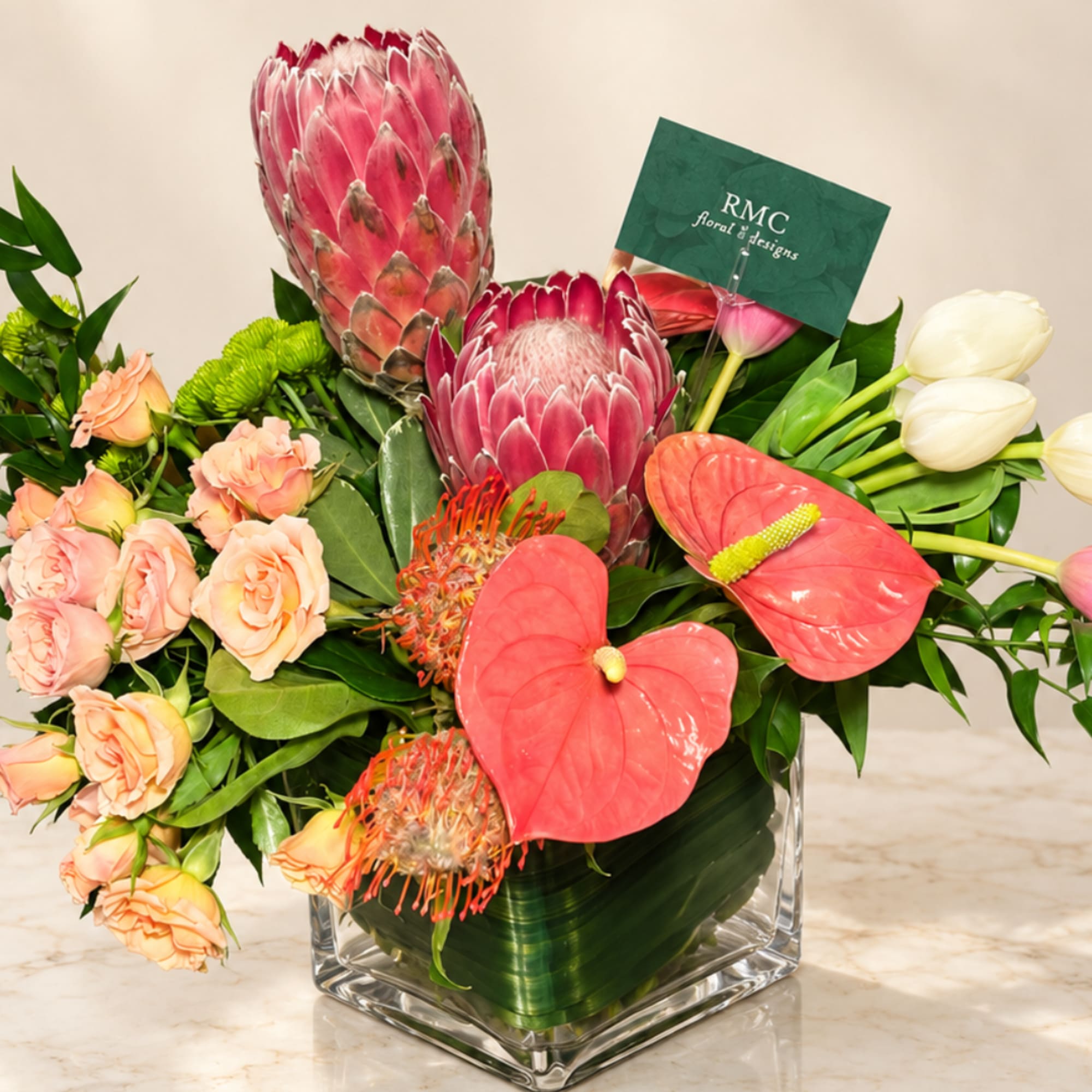 Tropical bouquet with pink protea, peach roses, red anthuriums, and white tulips in a glass vase