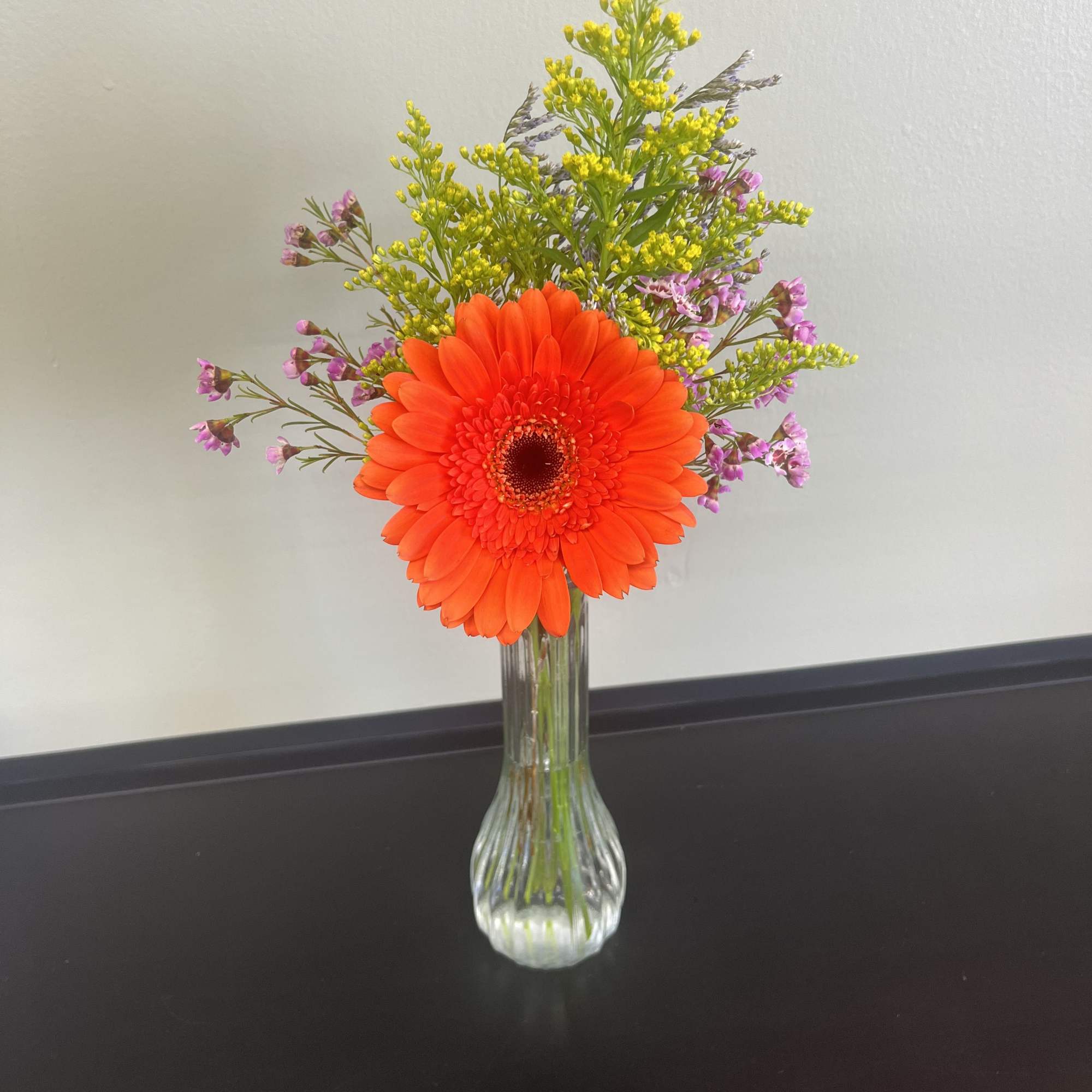 Orange gerbera daisy in a clear glass vase with small purple filler flowers