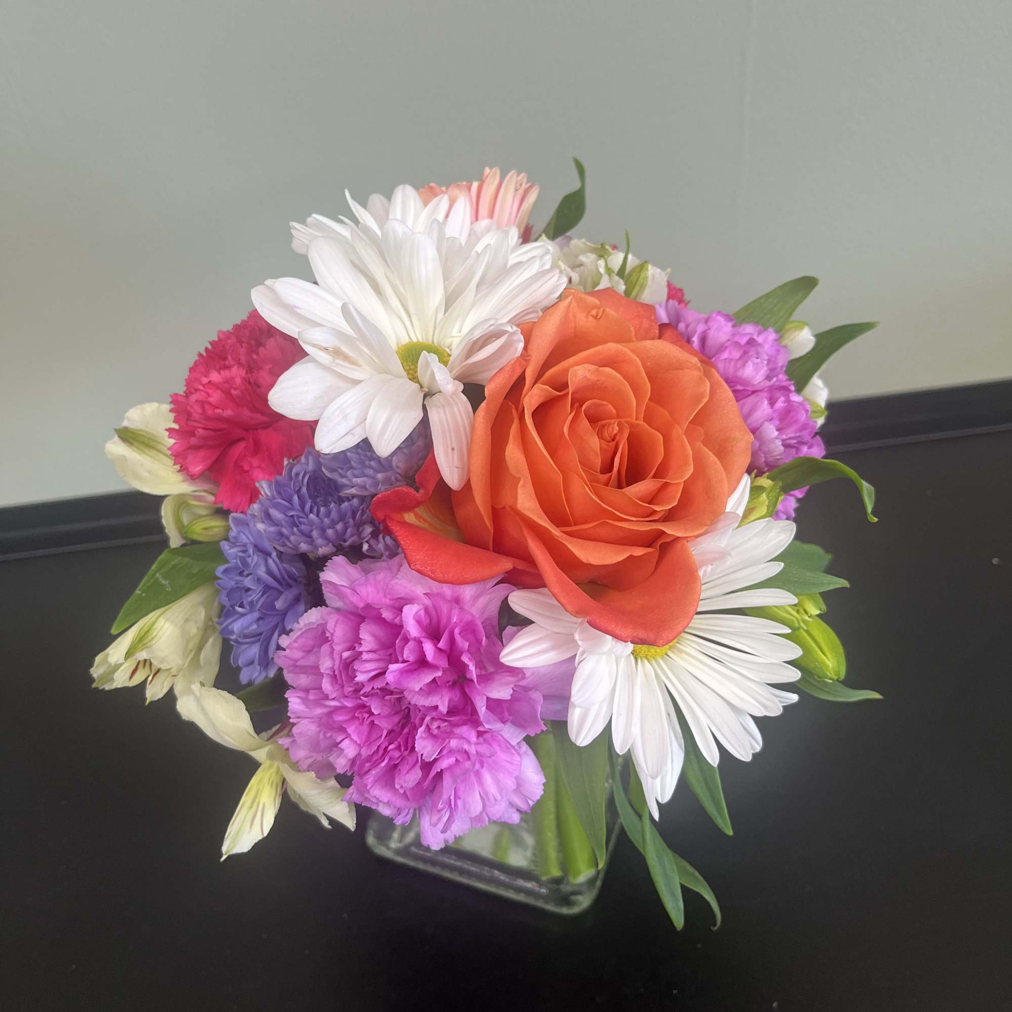 Mixed bouquet with an orange rose, white daisies, and pink and purple carnations in a glass vase