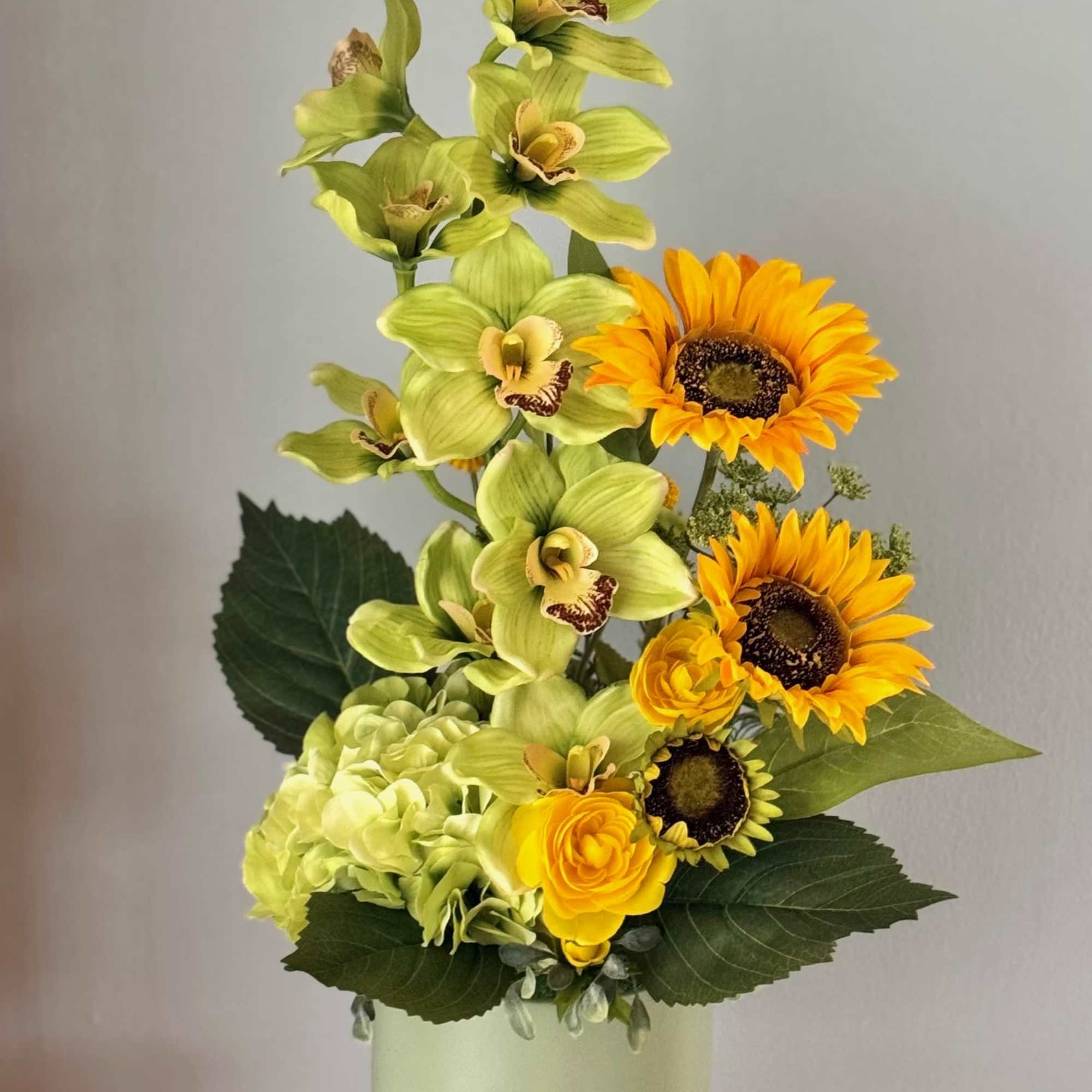 Tall arrangement of green orchids, sunflowers, and hydrangea in a pale vase