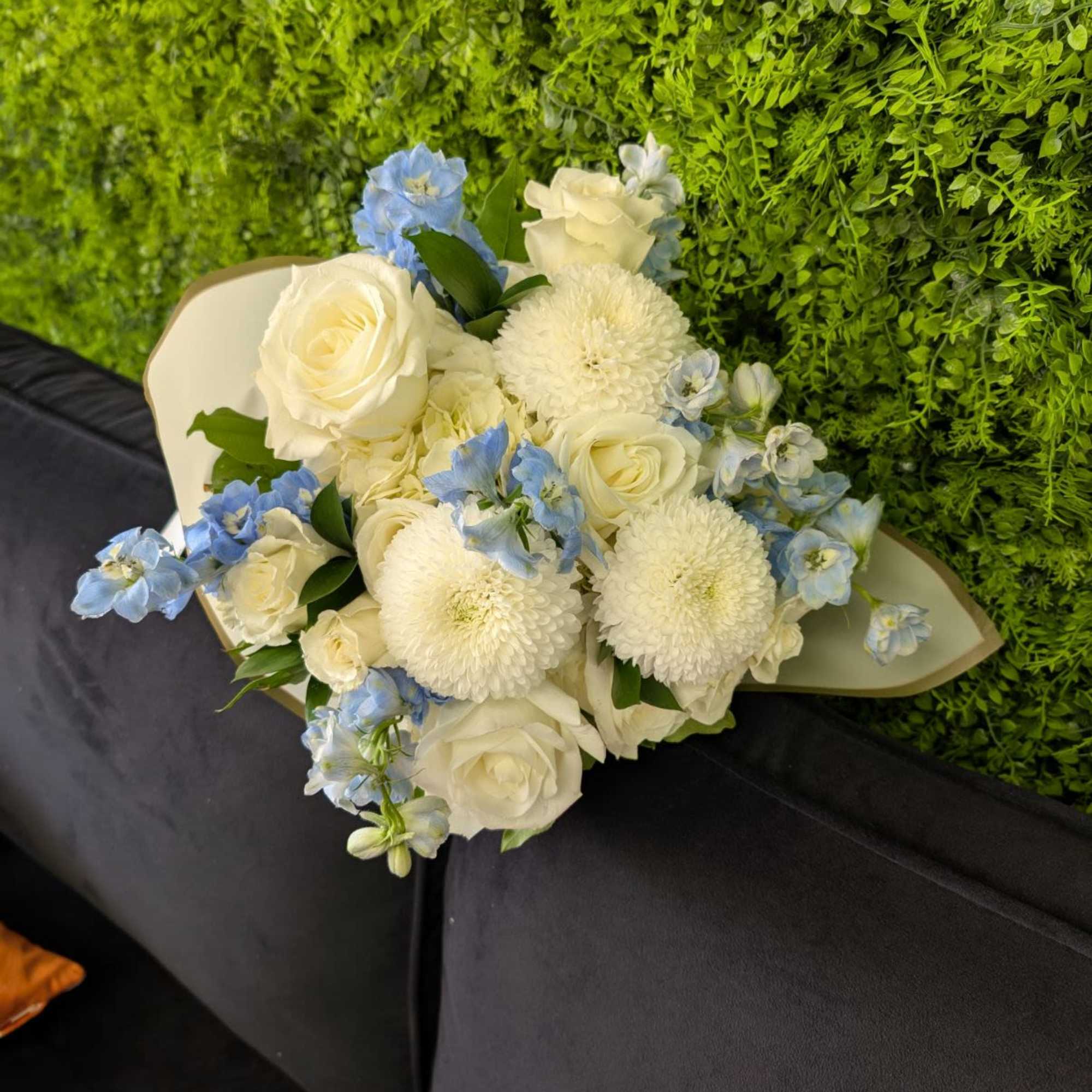 Hand-tied bouquet of white roses, white mums, and pale blue flowers resting on a dark sofa.