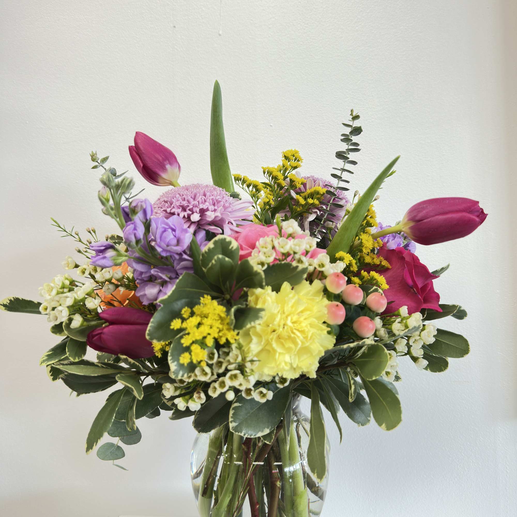 Mixed bouquet of pink tulips, yellow carnation, and purple blooms in a clear glass vase.