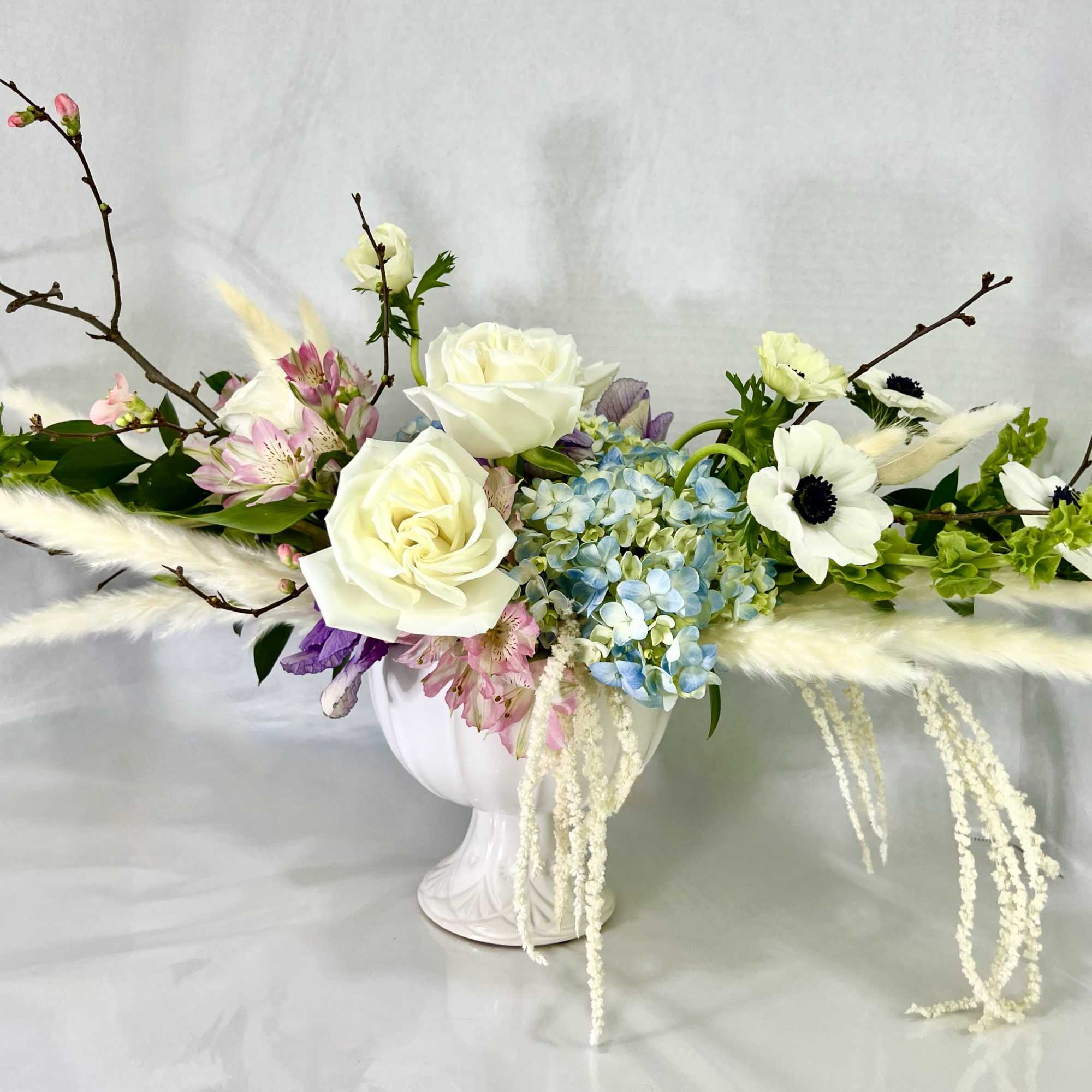 White roses and blue hydrangeas arranged in a white vase with branches.