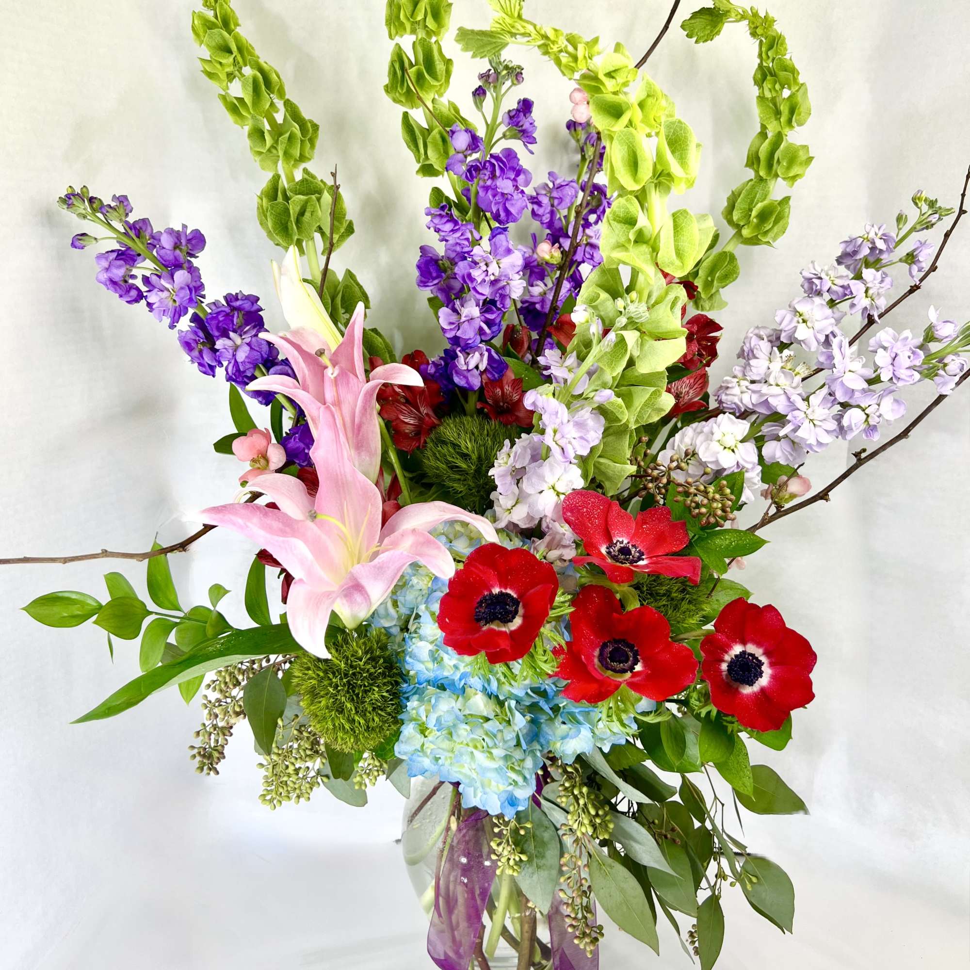 Tall bouquet of pink lilies, red anemones, and purple blooms in a glass vase