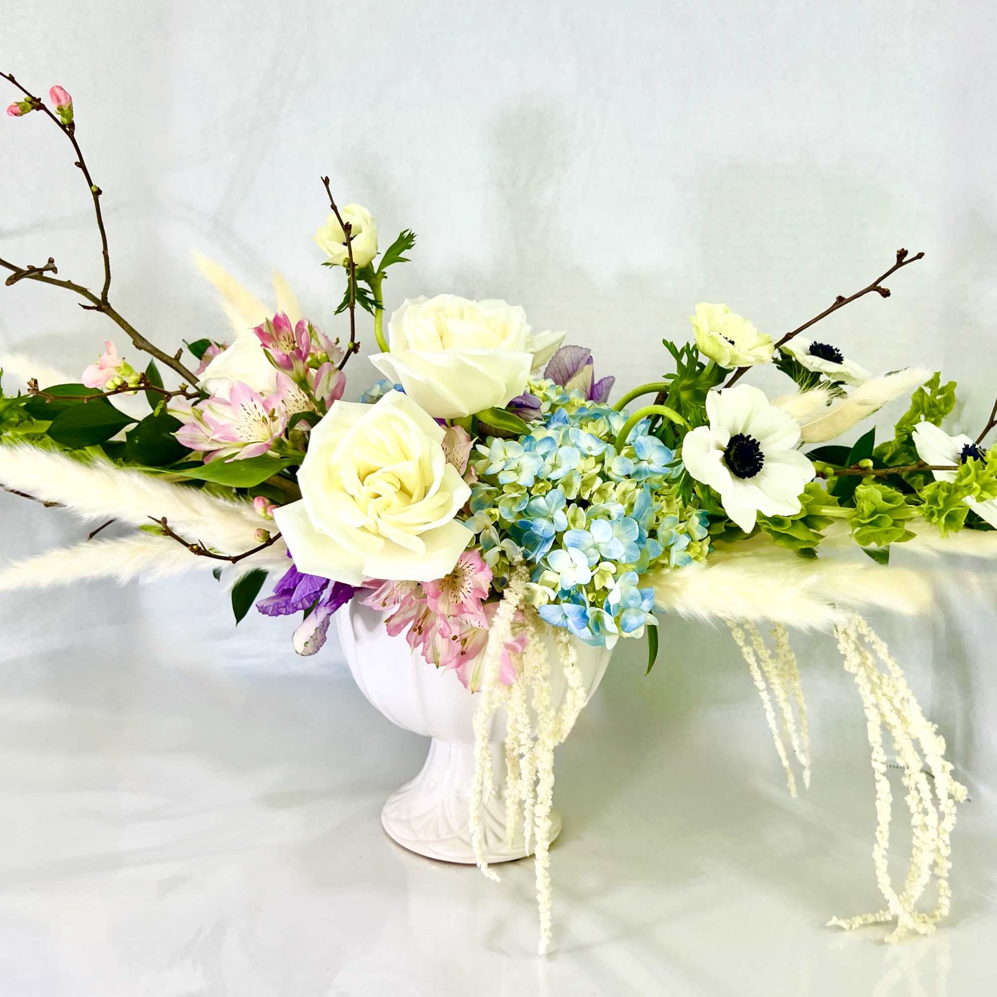 White roses and blue hydrangeas in a white vase with branches and feathers