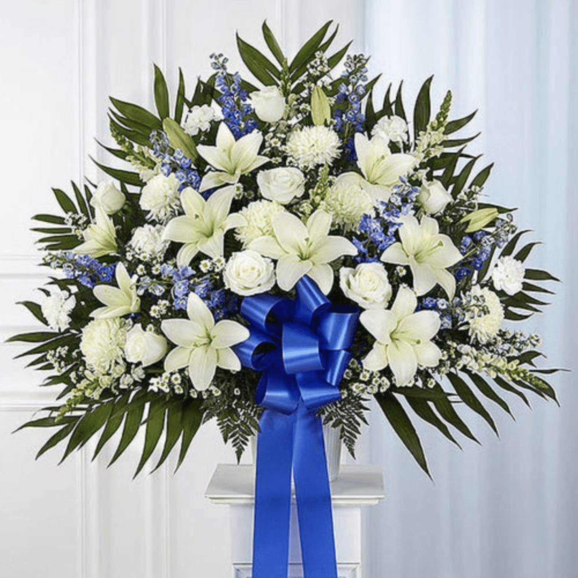 White lilies and roses with blue flowers in a standing spray with a blue ribbon