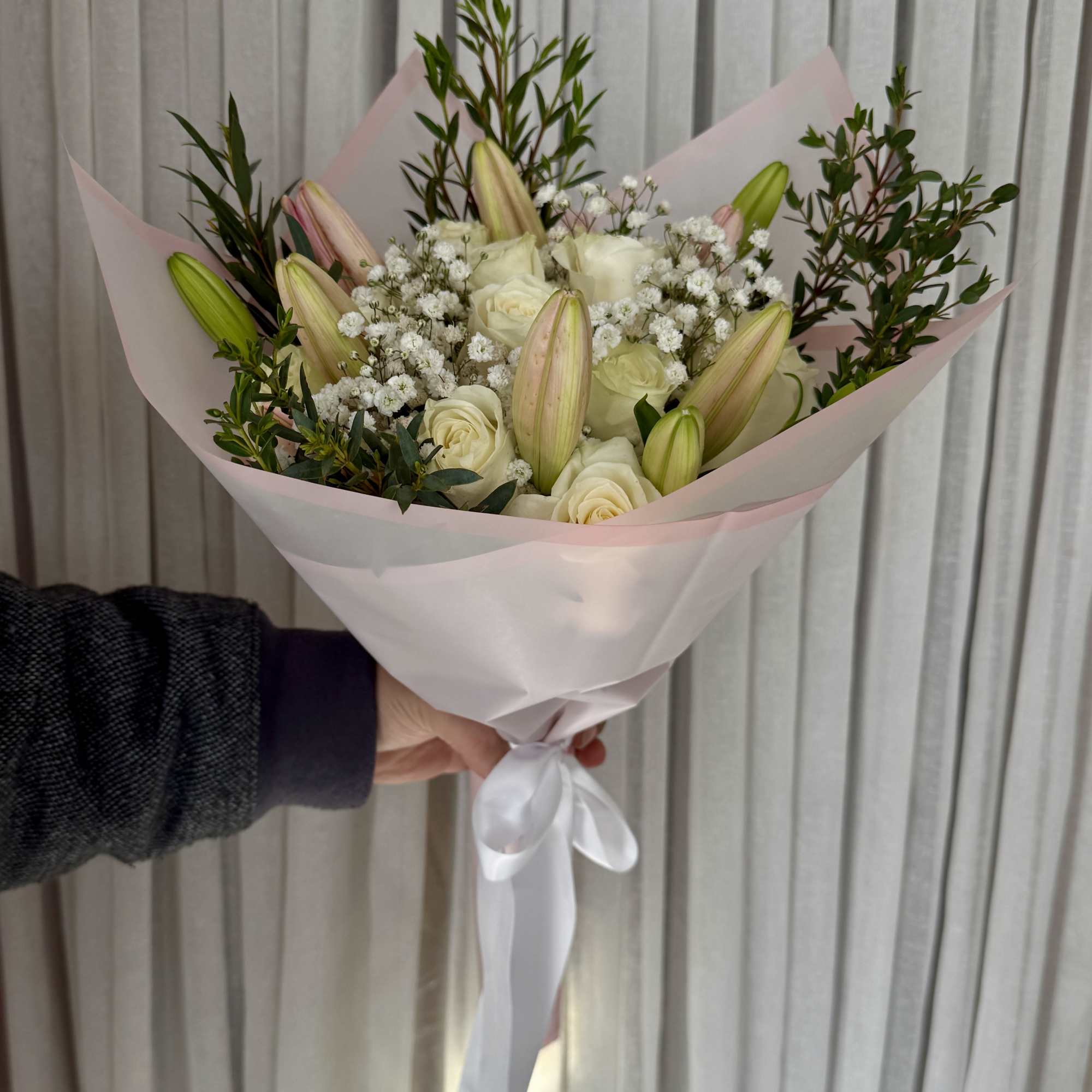 Handheld bouquet of white roses and lily buds wrapped in pale pink paper