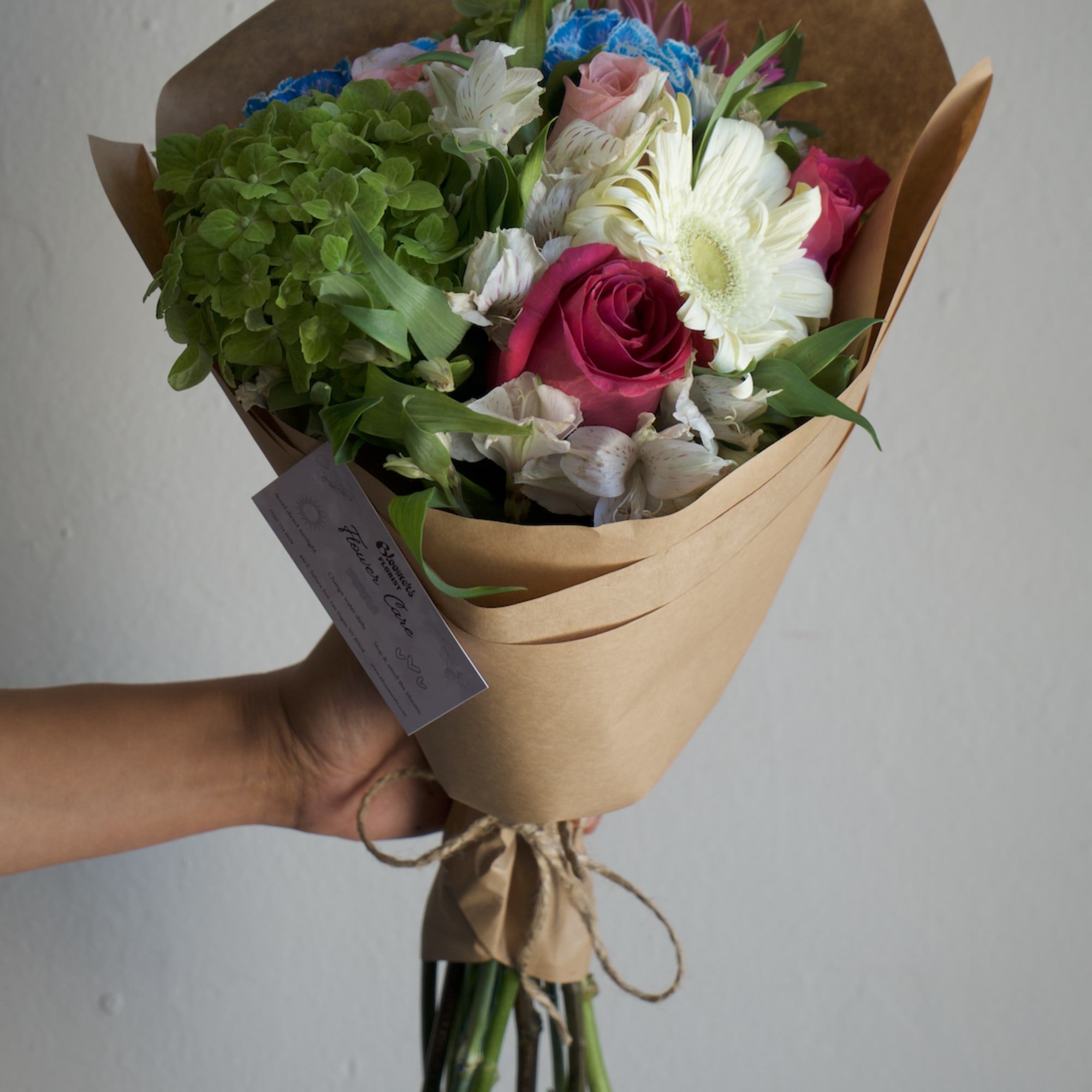 Handheld bouquet with pink roses, white gerbera daisies, and green hydrangea in brown paper
