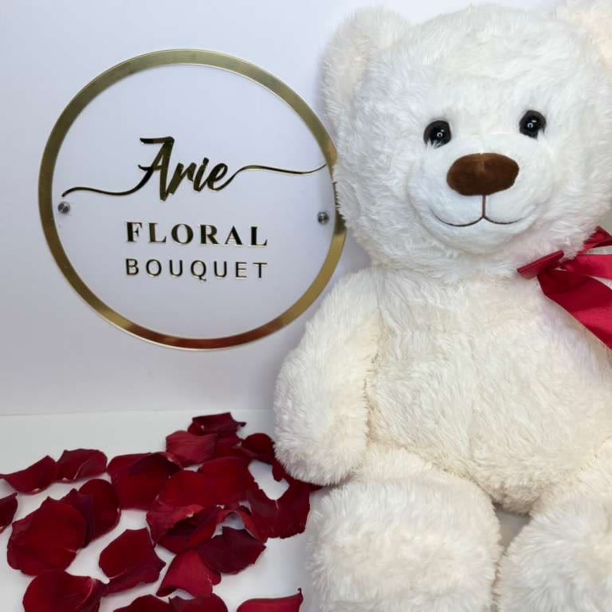 White teddy bear with a red ribbon beside rose petals and a floral bouquet sign