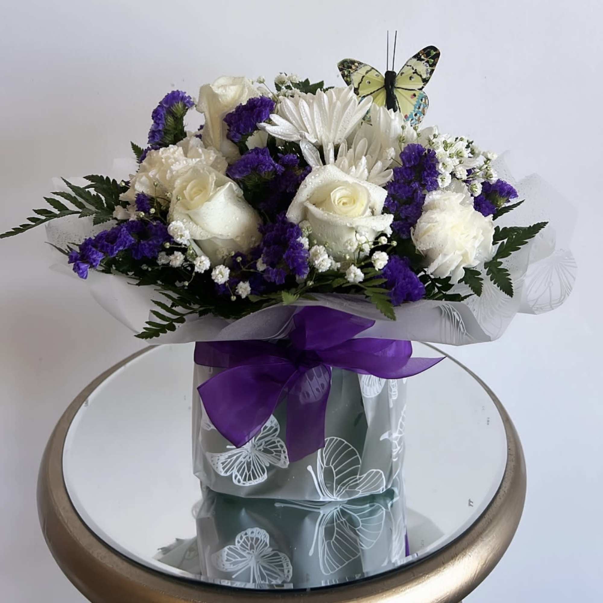 White rose bouquet with purple filler flowers and a butterfly topper in a wrapped container