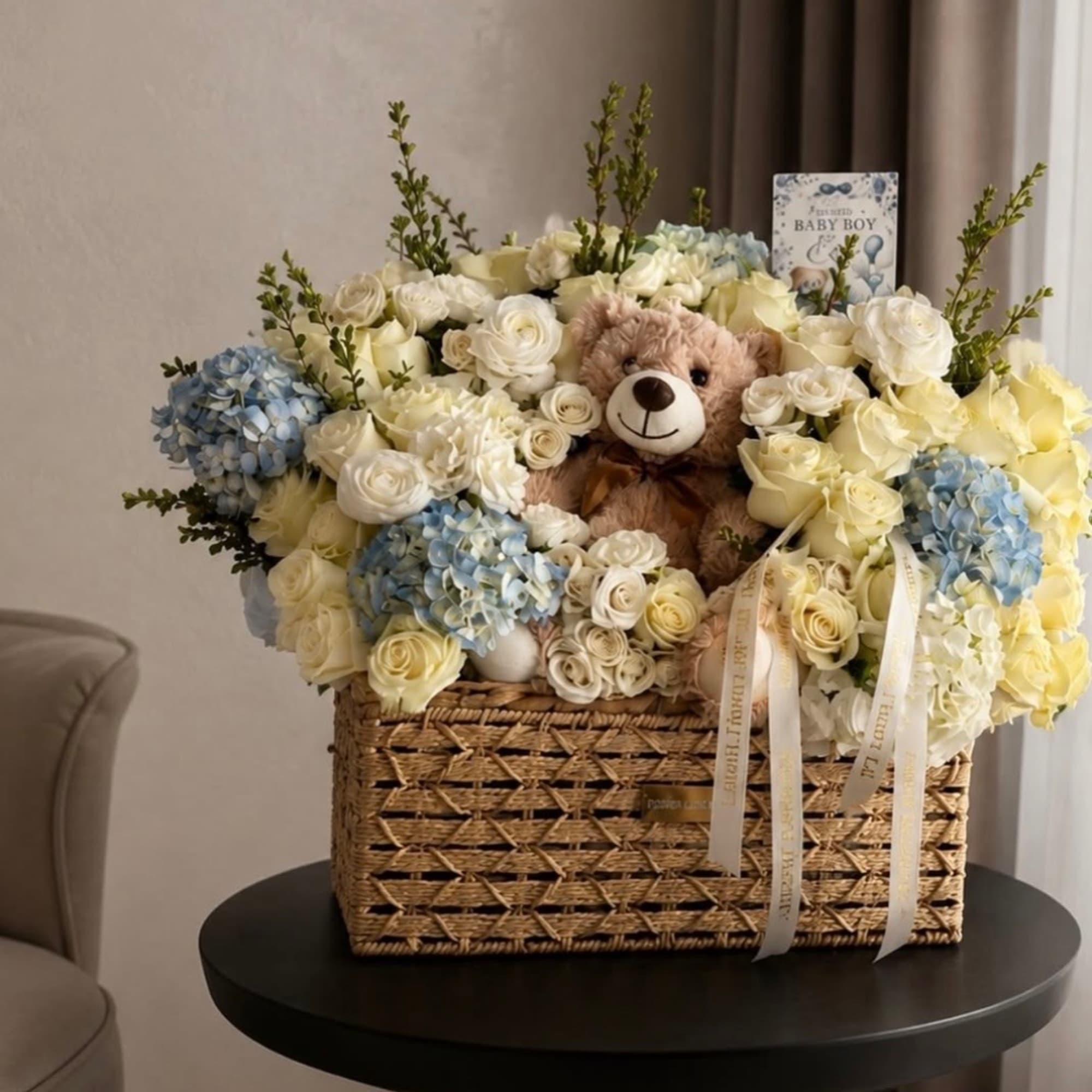 Basket arrangement of white and blue flowers with a teddy bear and baby boy card