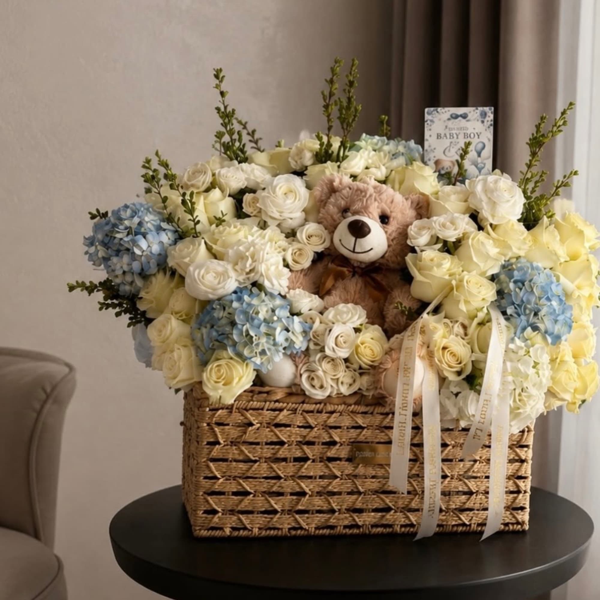 Basket arrangement of white and blue flowers with a teddy bear and baby boy card