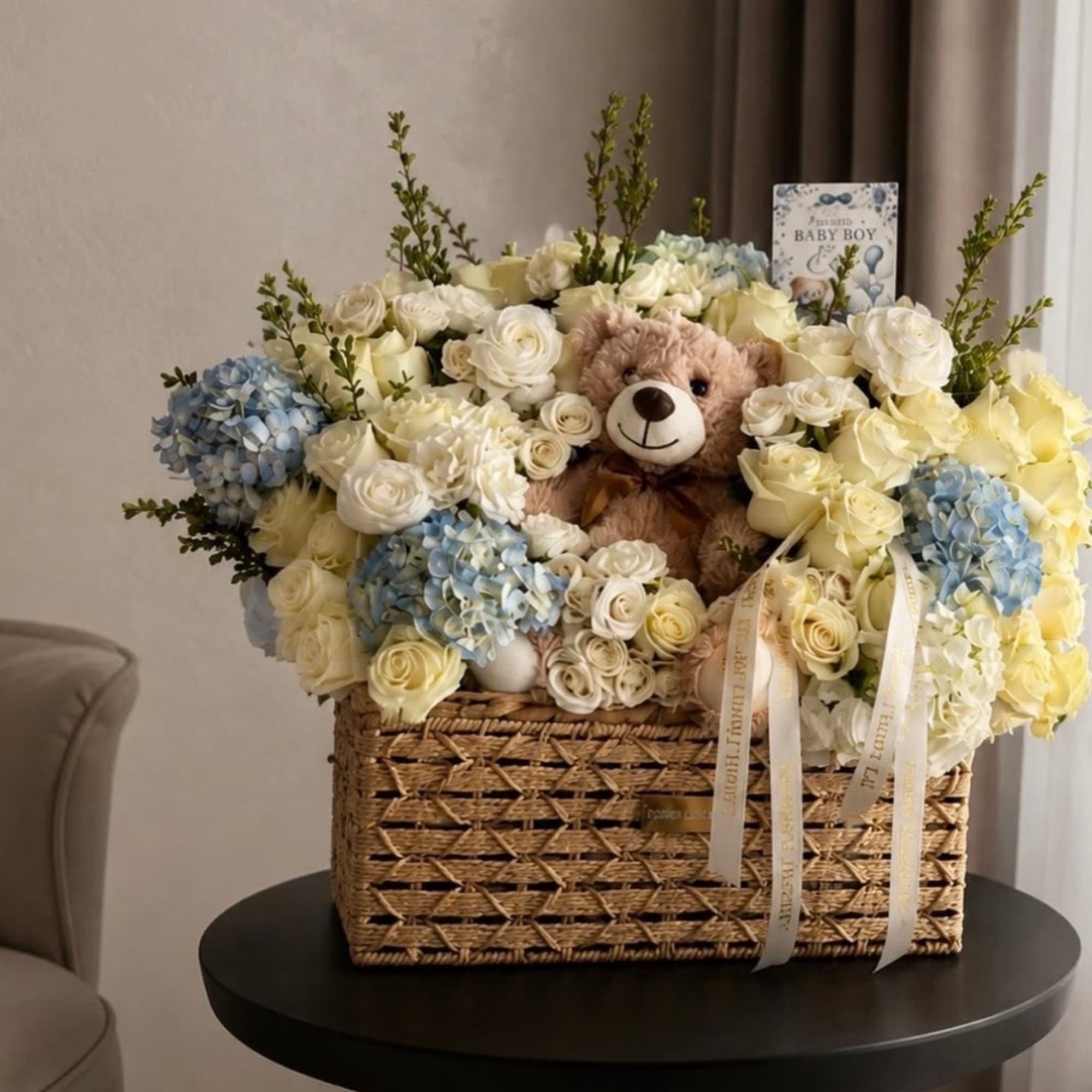 Basket arrangement of white and blue flowers with a teddy bear and baby card
