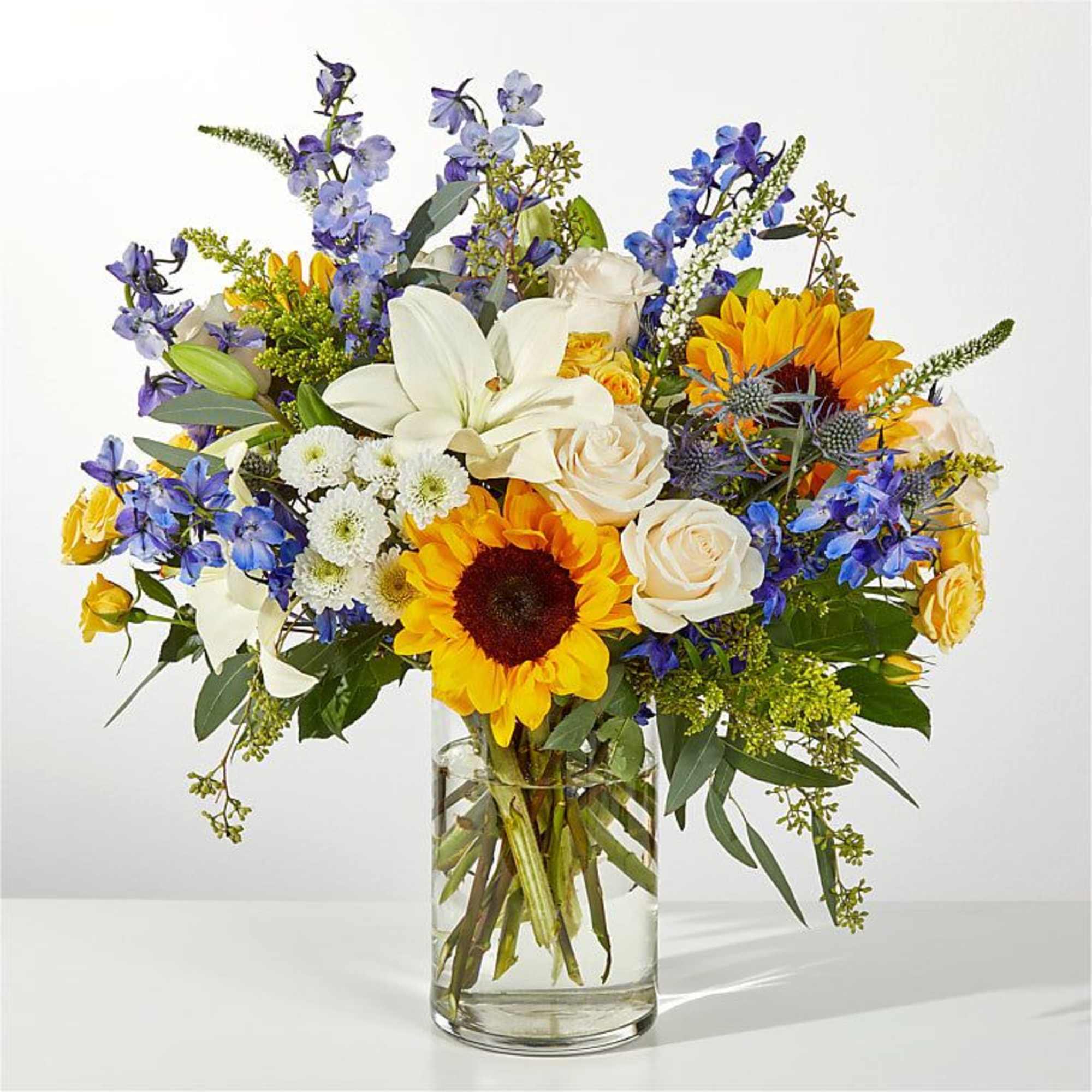 Mixed bouquet with sunflowers, white lilies, and blue flowers in a glass vase