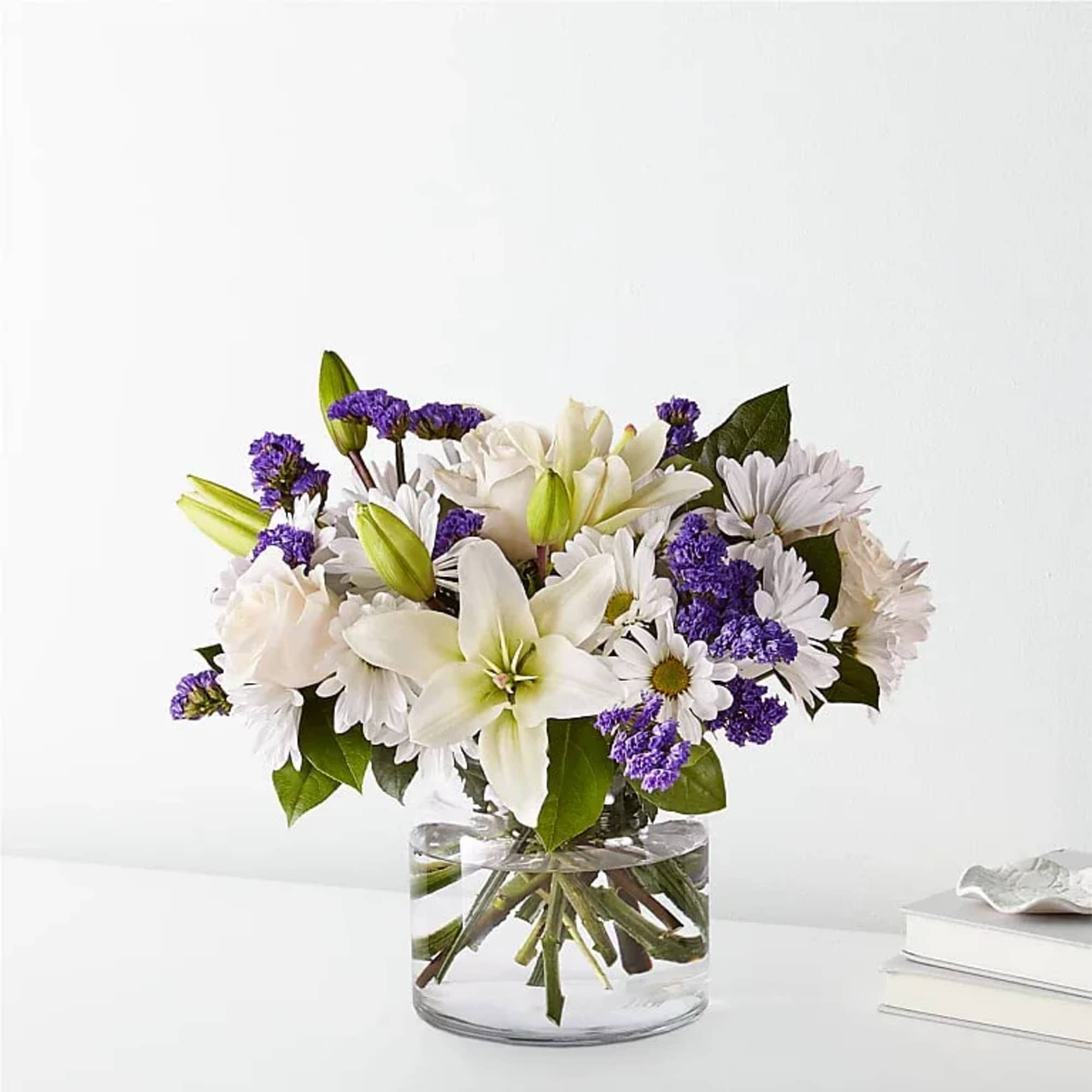 White lilies and daisies with purple filler in a clear glass vase