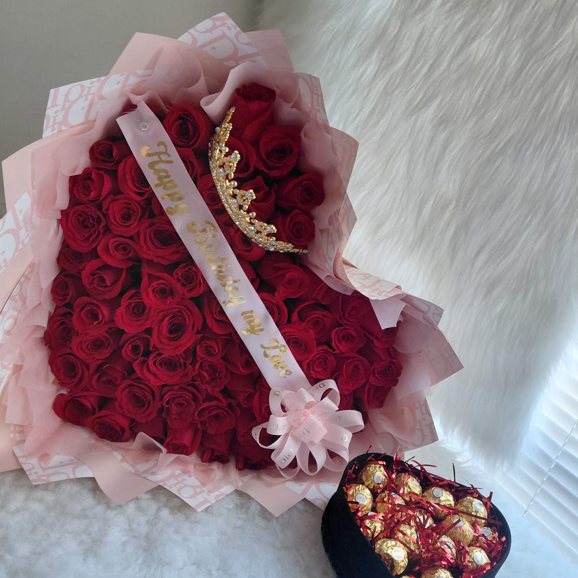Bouquet of red roses with a birthday ribbon and tiara, plus a box of chocolates.