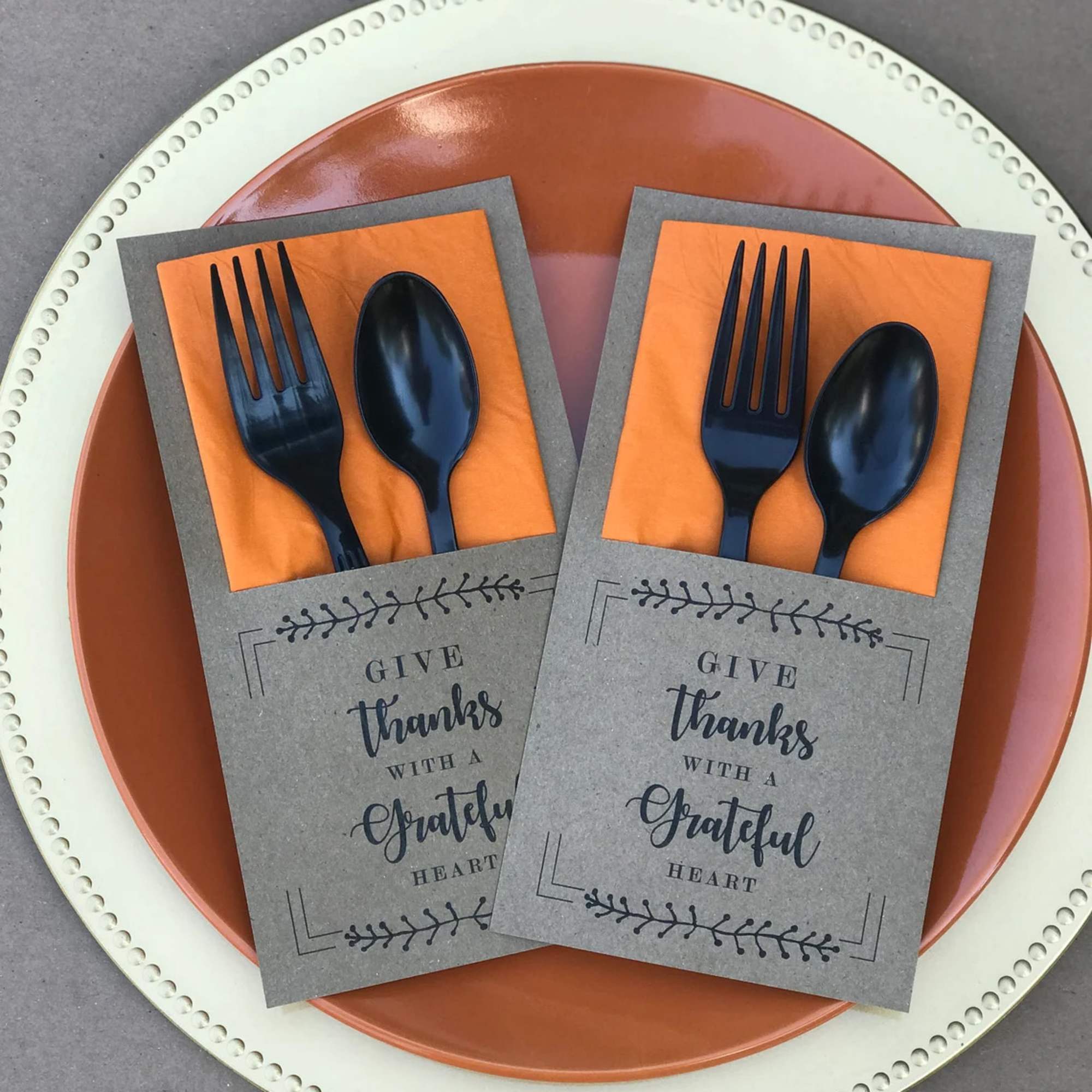 Two gratitude-themed napkin holders with black plastic forks and spoons on layered plates