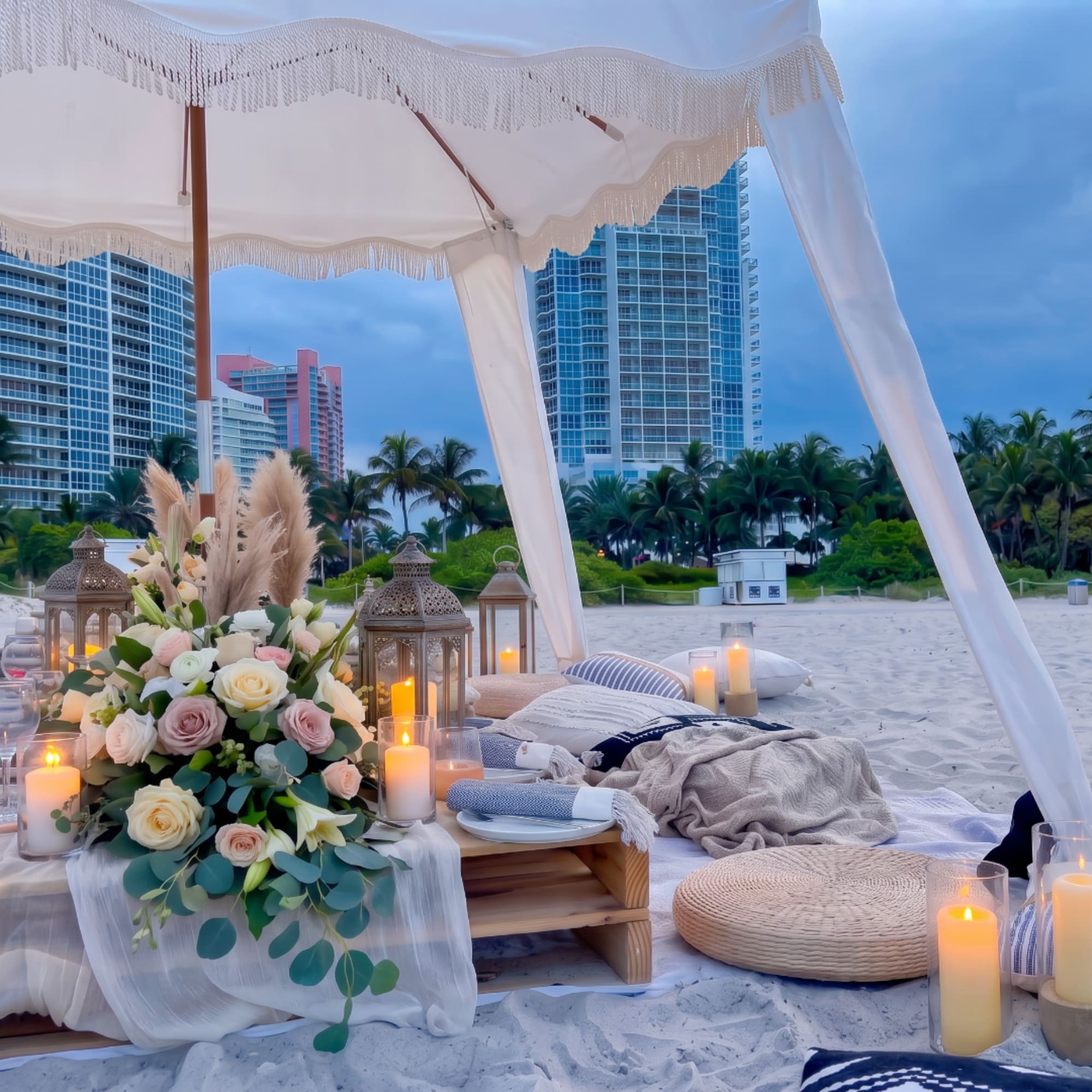 Beach picnic setup with a pastel rose arrangement, lanterns, and candles under a canopy