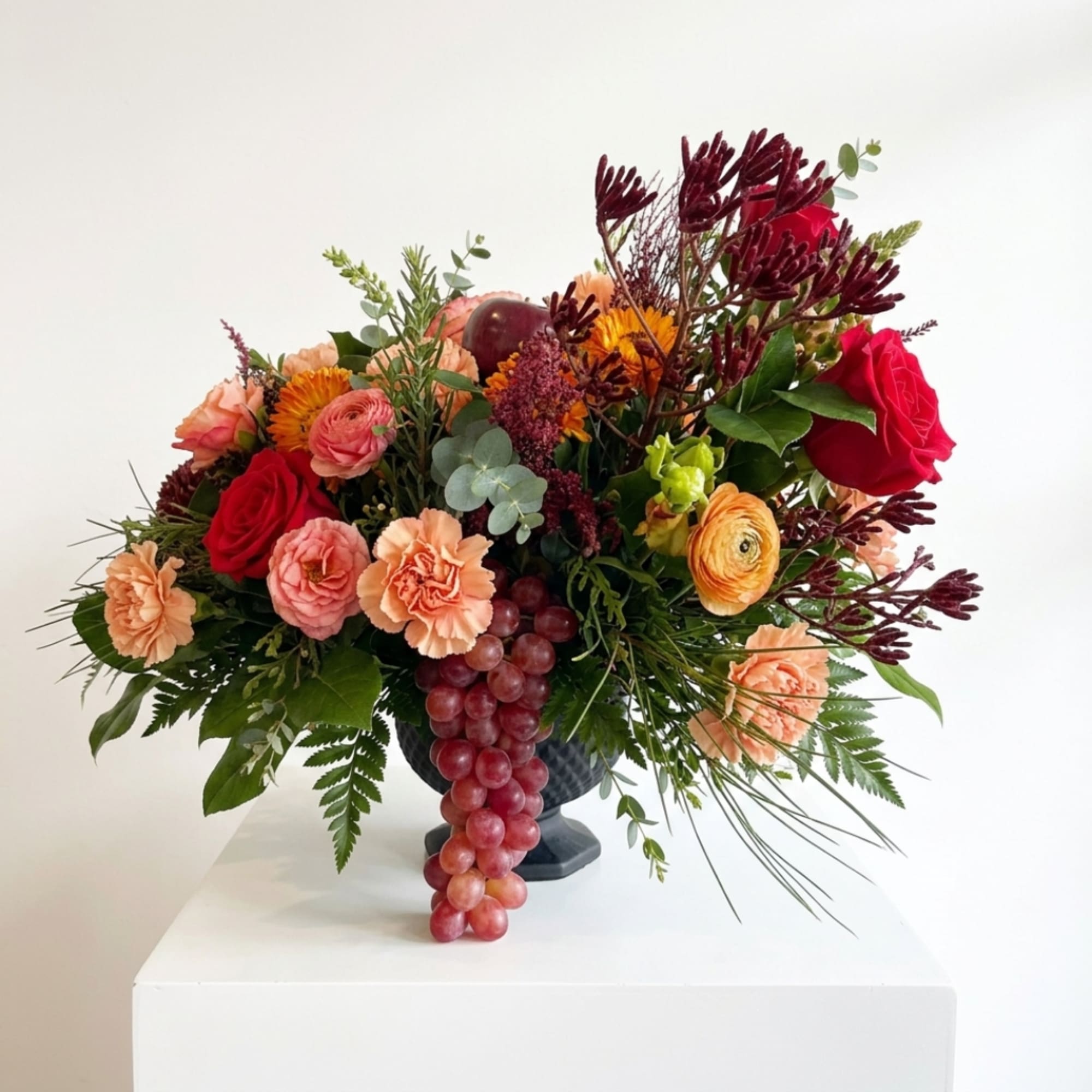 Mixed bouquet in a dark vase with red roses, peach carnations, and hanging grapes
