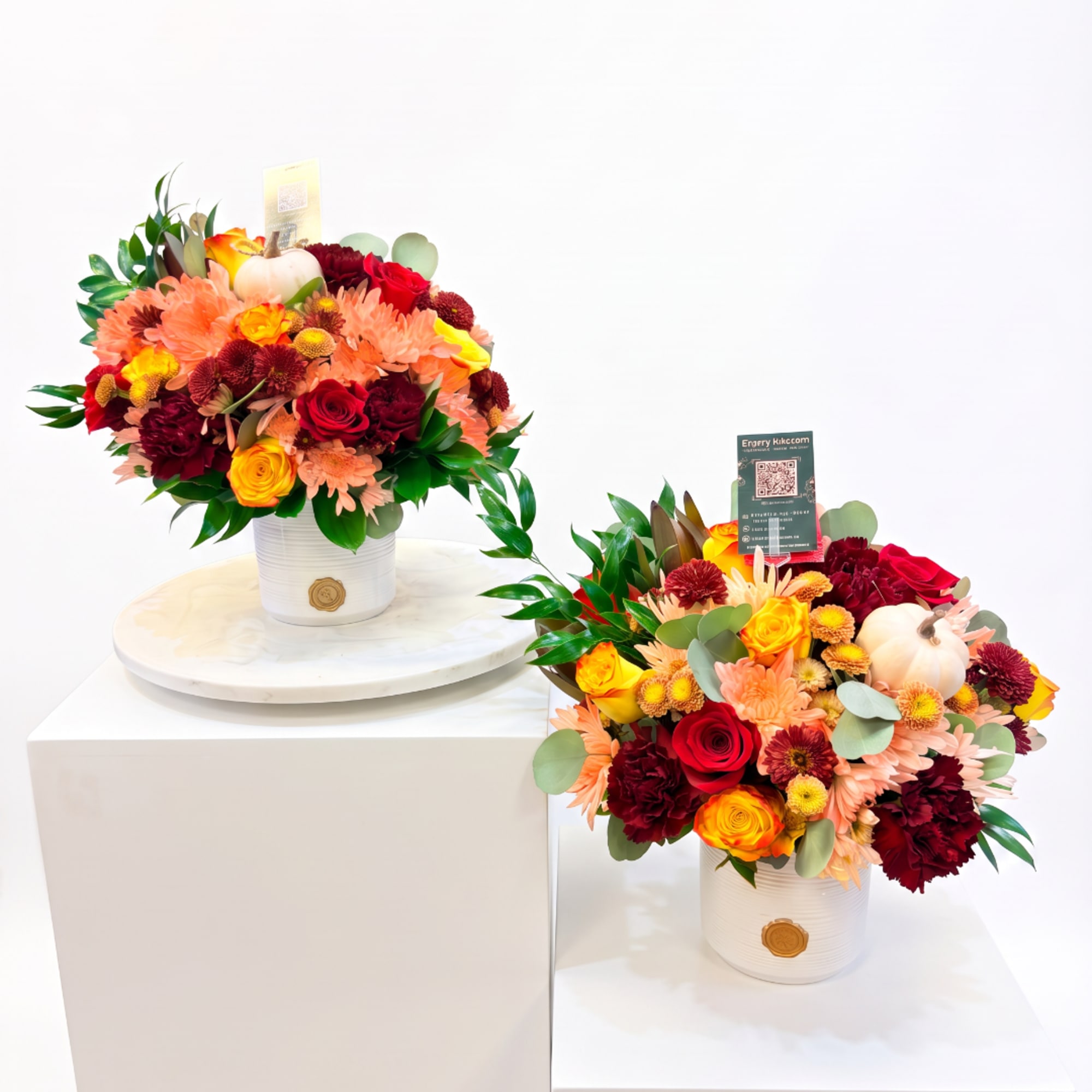 Two colorful flower arrangements in white containers with small pumpkins