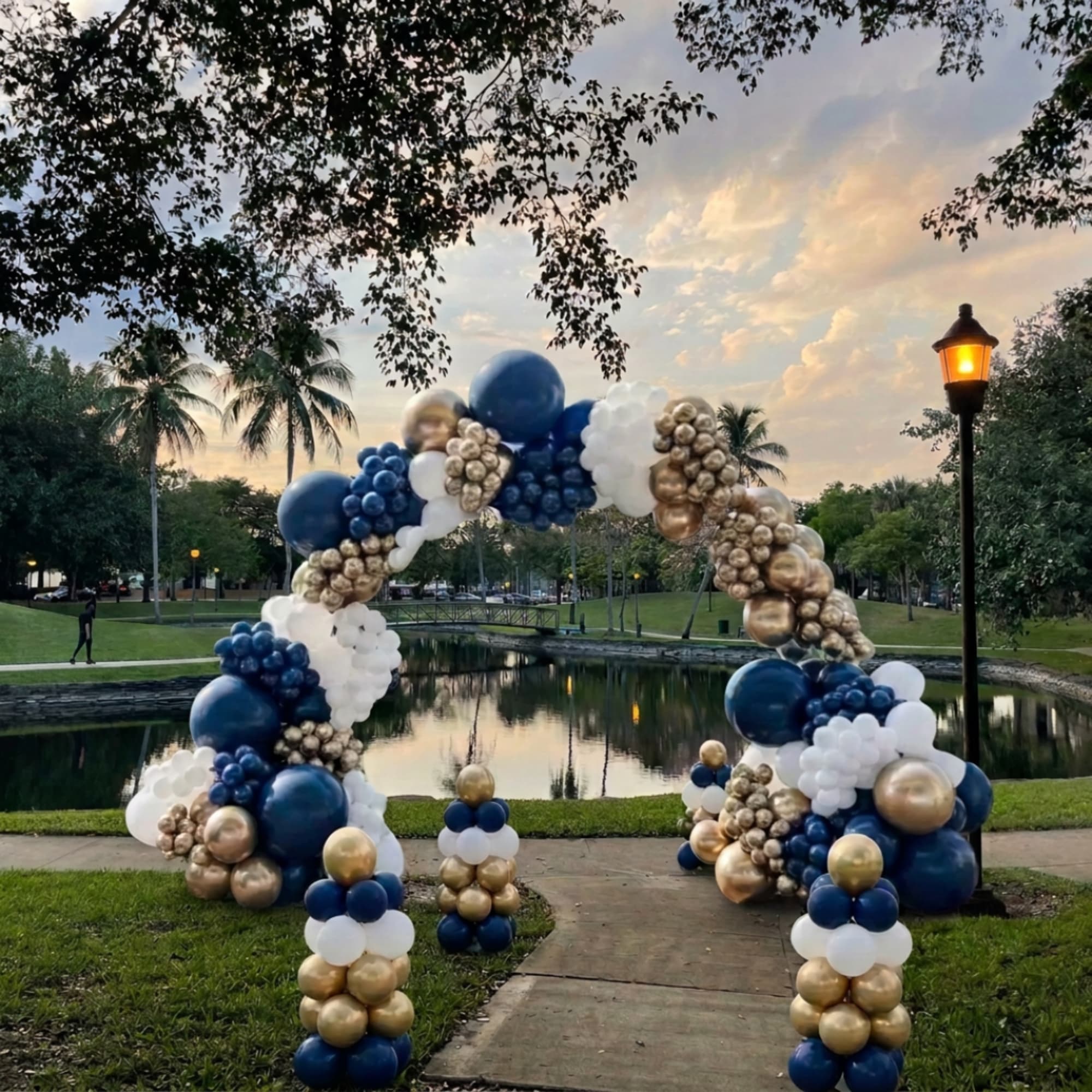 Large balloon arch in blue, white, and gold beside a park pond at sunset