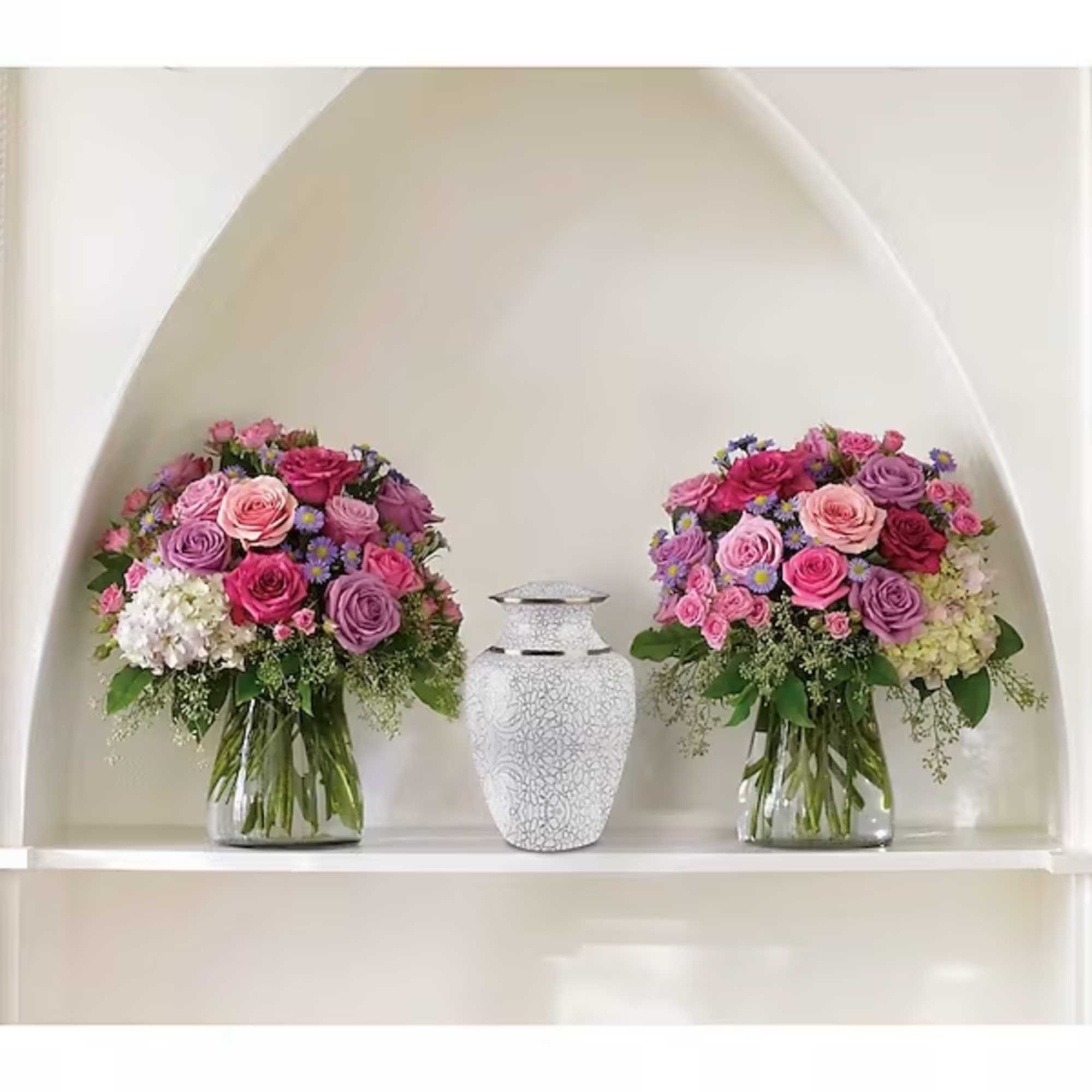 Two pink and purple rose arrangements in glass vases with a white urn between them