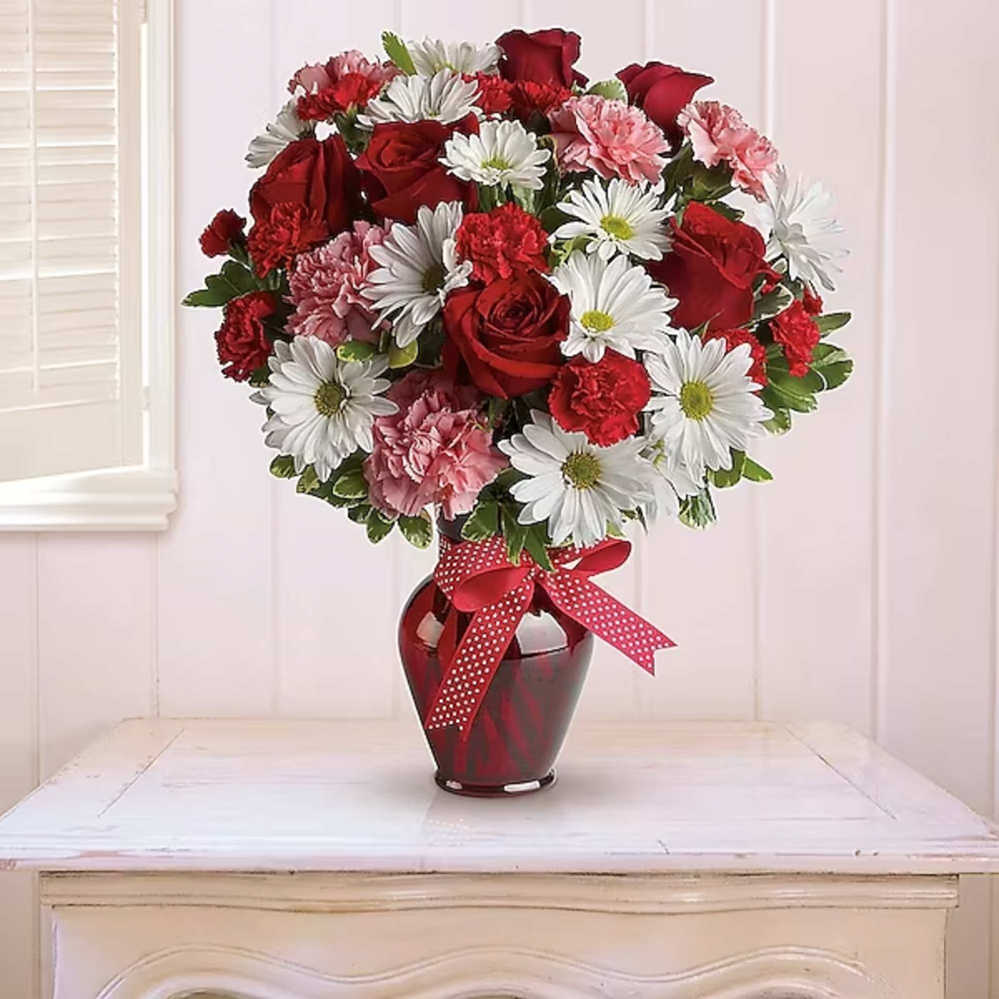 Red roses, pink carnations, and white daisies in a red vase with a ribbon.