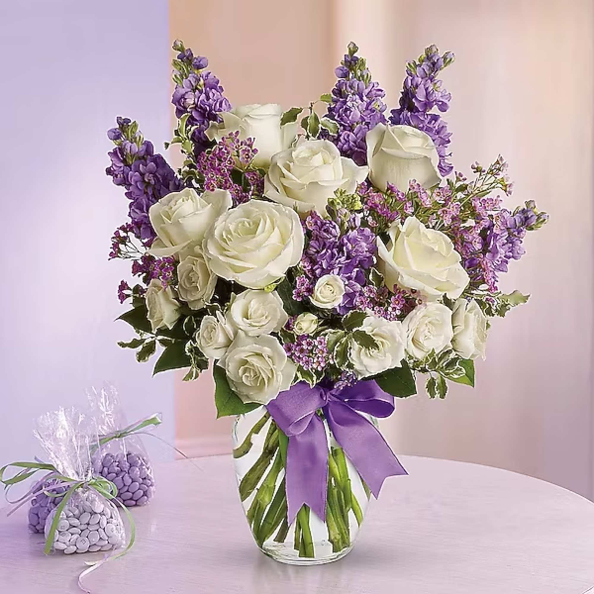 White roses and purple flowers arranged in a glass vase with a purple ribbon.