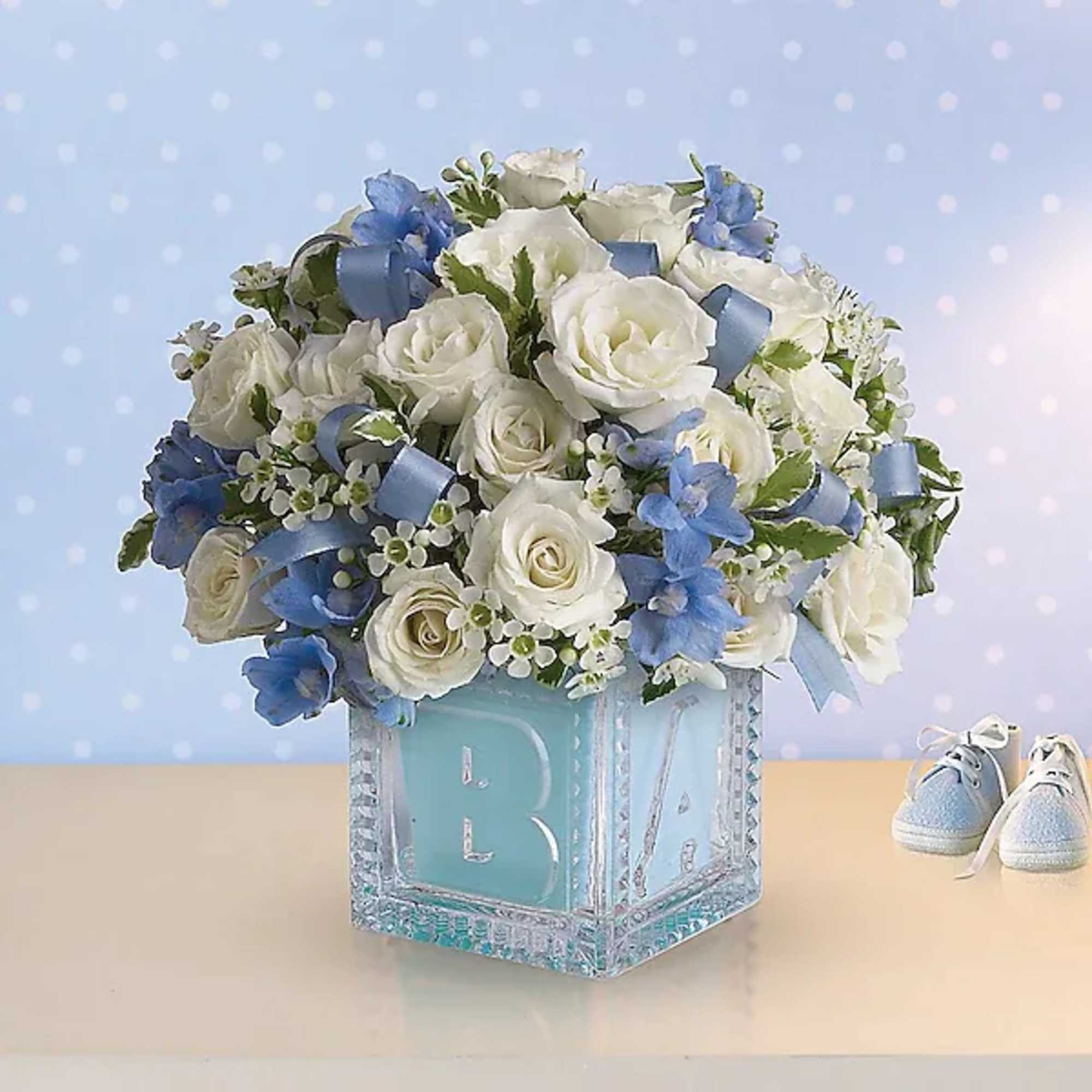 White roses and blue flowers in a glass block vase with baby shoes nearby