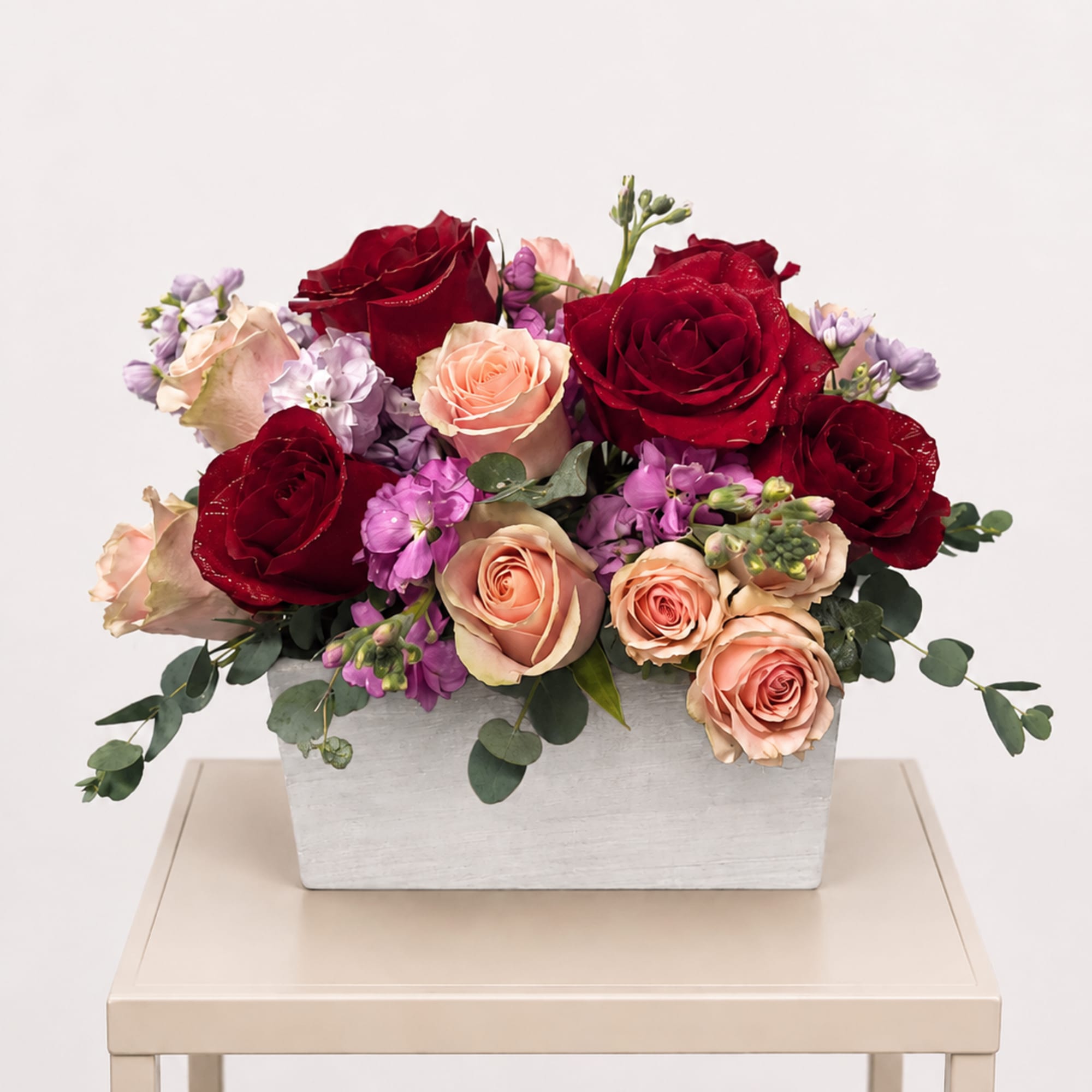 Red and blush roses with purple blooms in a white rectangular vase