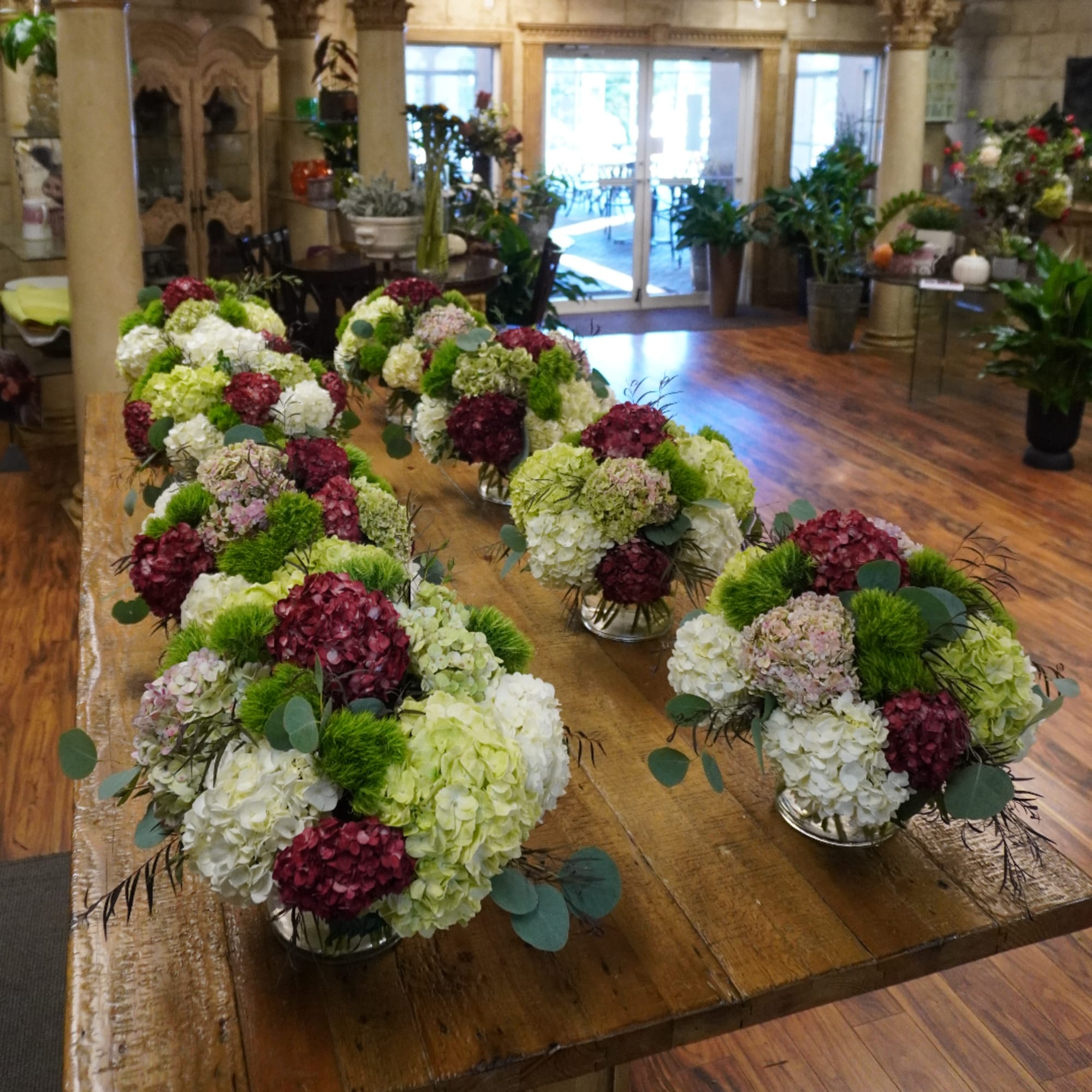 Several low floral centerpieces with hydrangeas in white, green, and burgundy on a wooden table.
