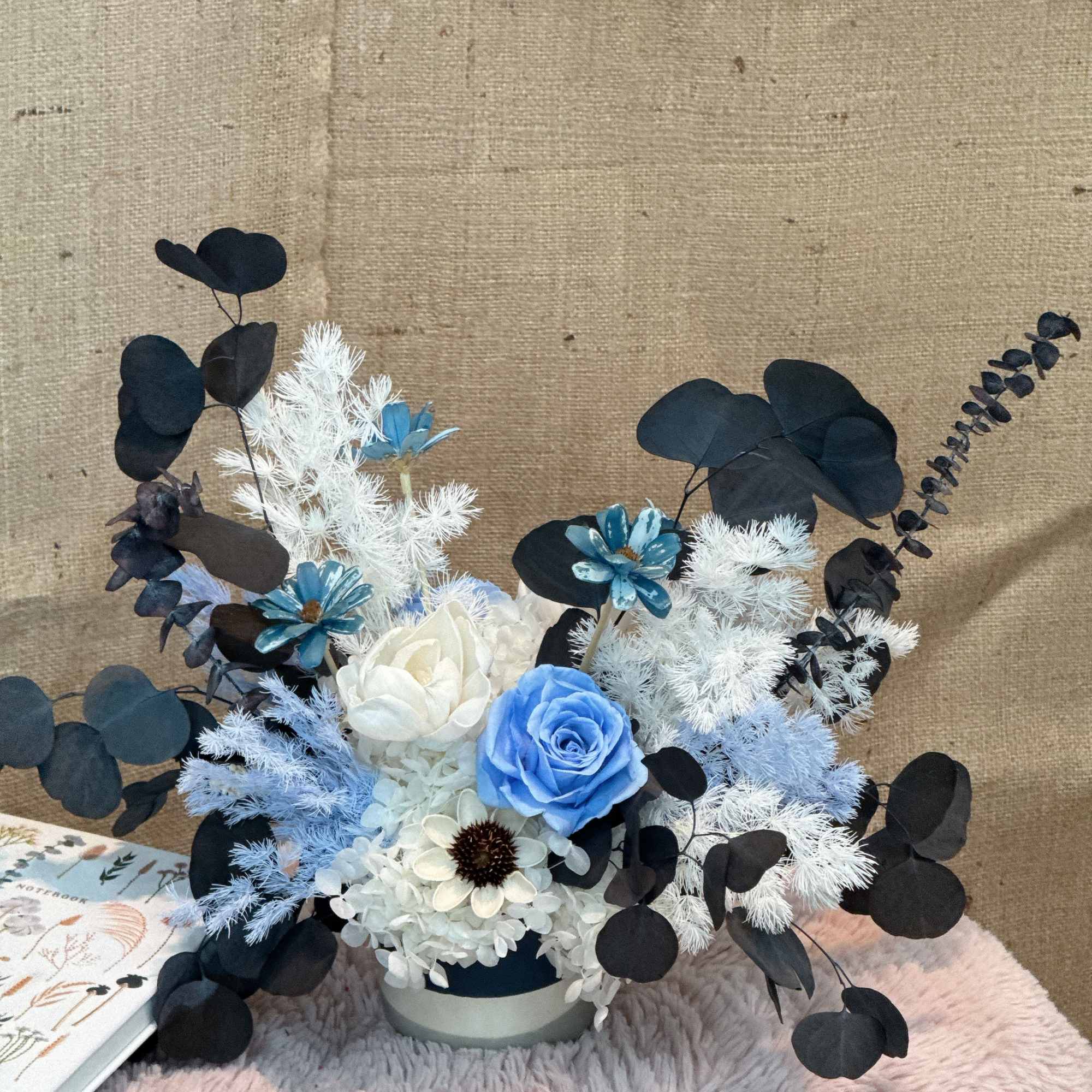 Modern arrangement of blue and white flowers with dark foliage in a white pot on a soft surface