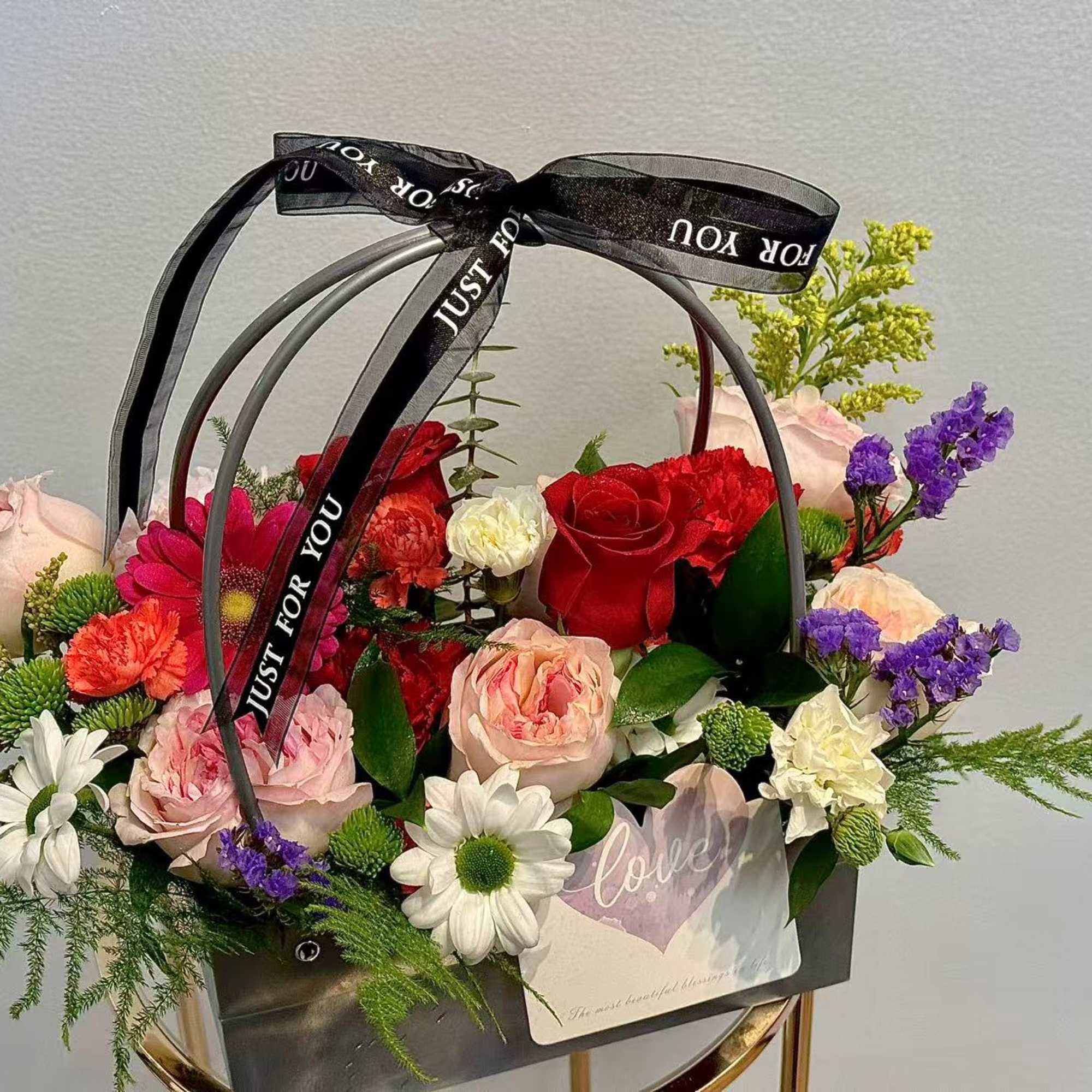Mixed flower arrangement in a black handled basket with a ribbon bow