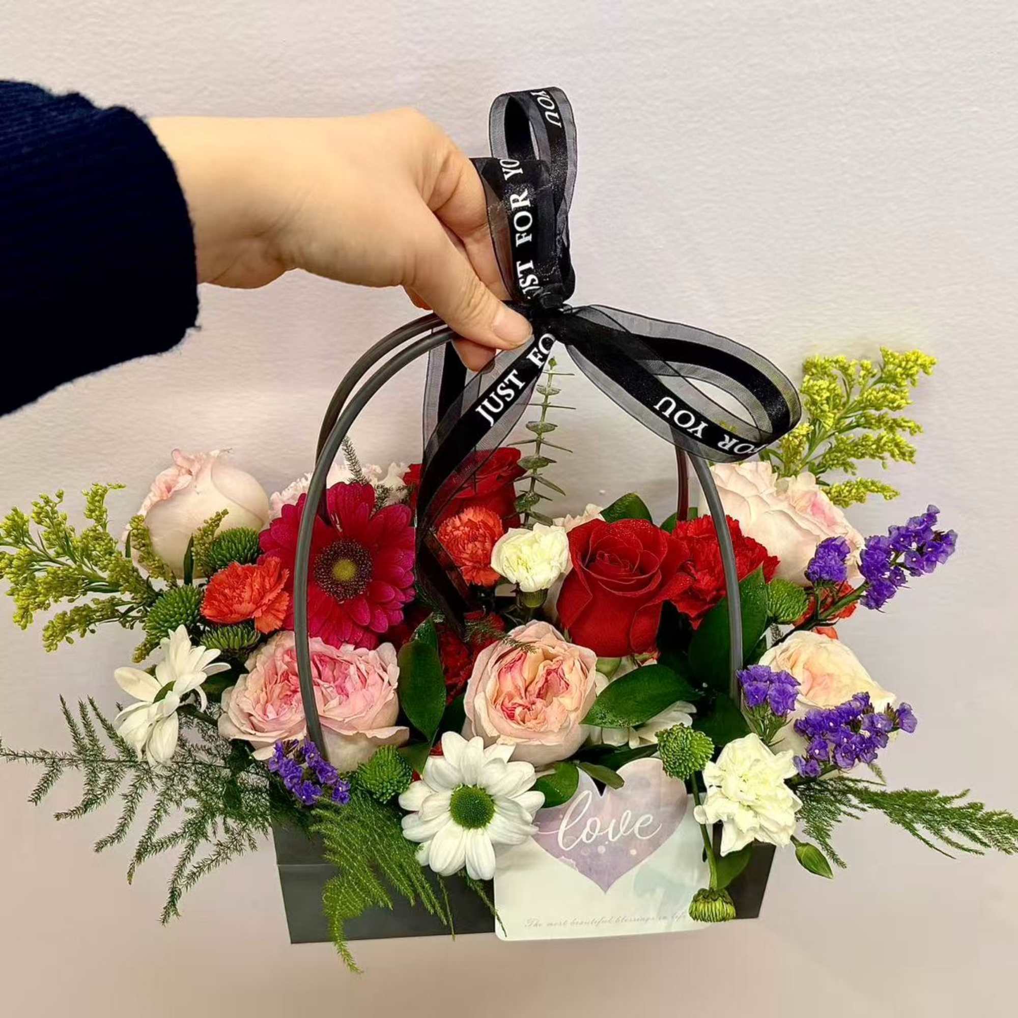 Handheld basket arrangement of mixed roses, daisies, and carnations with a black ribbon