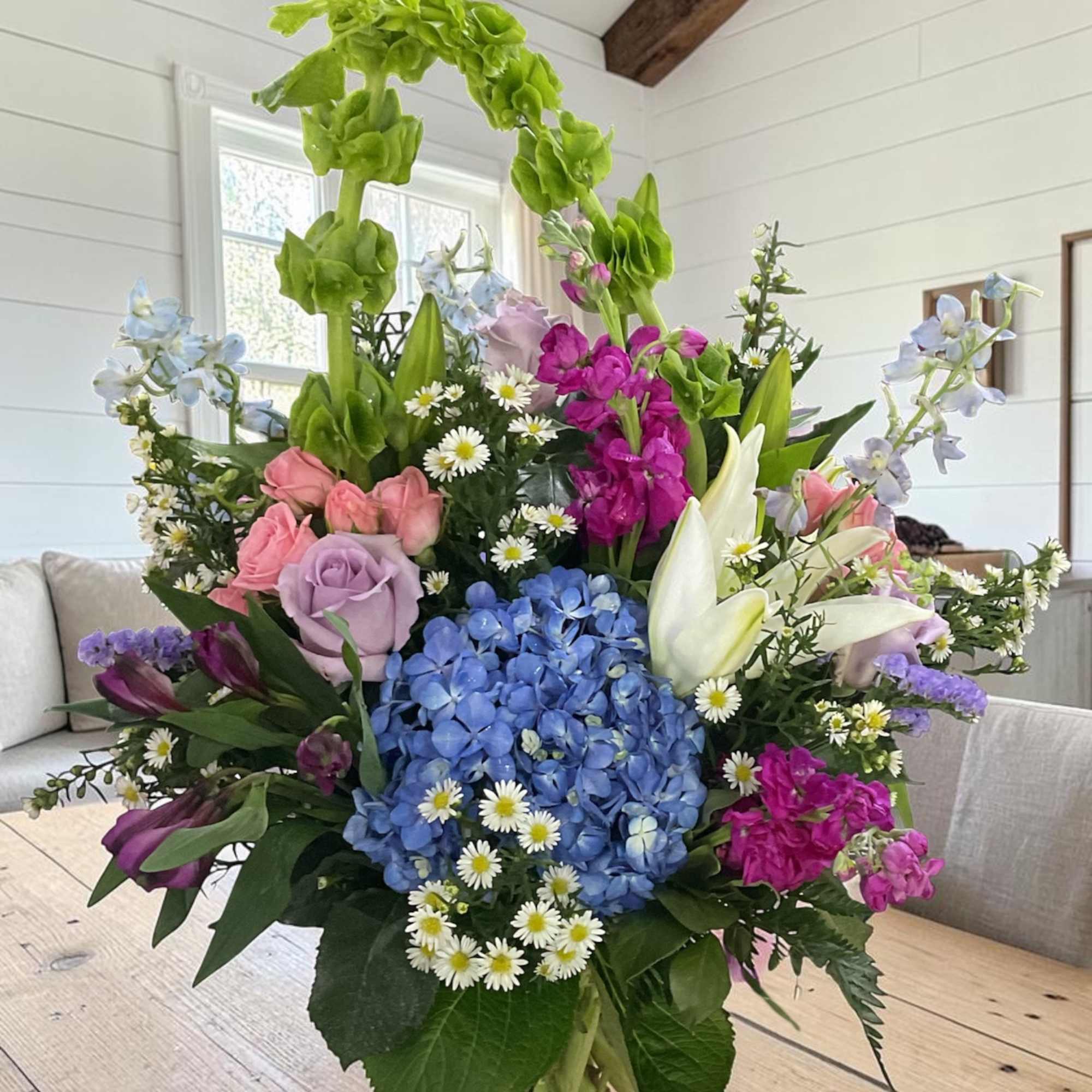 Mixed bouquet in a clear glass vase with blue hydrangea, roses, lilies, and tall green blooms