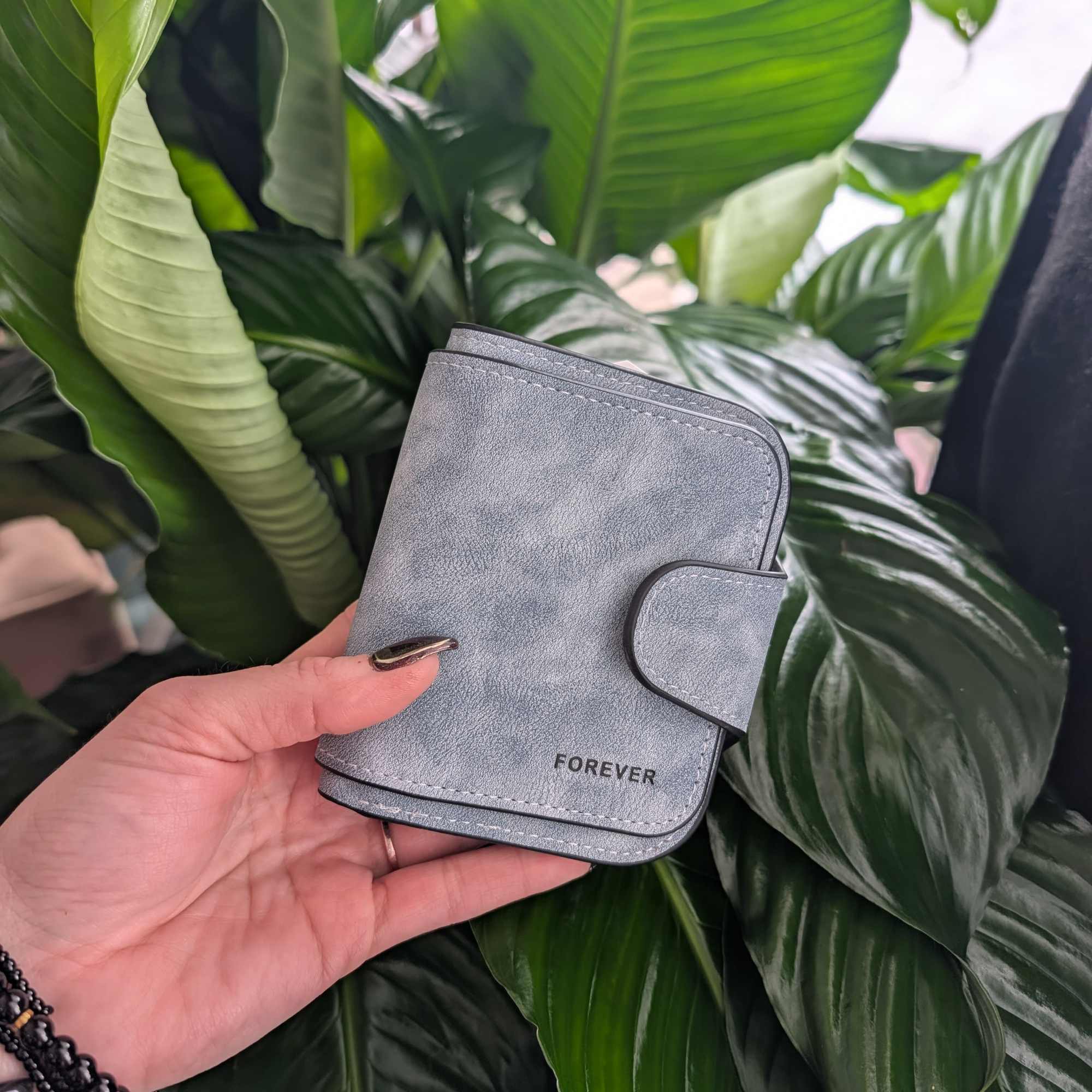 Hand holding a gray wallet in front of large green plant leaves.