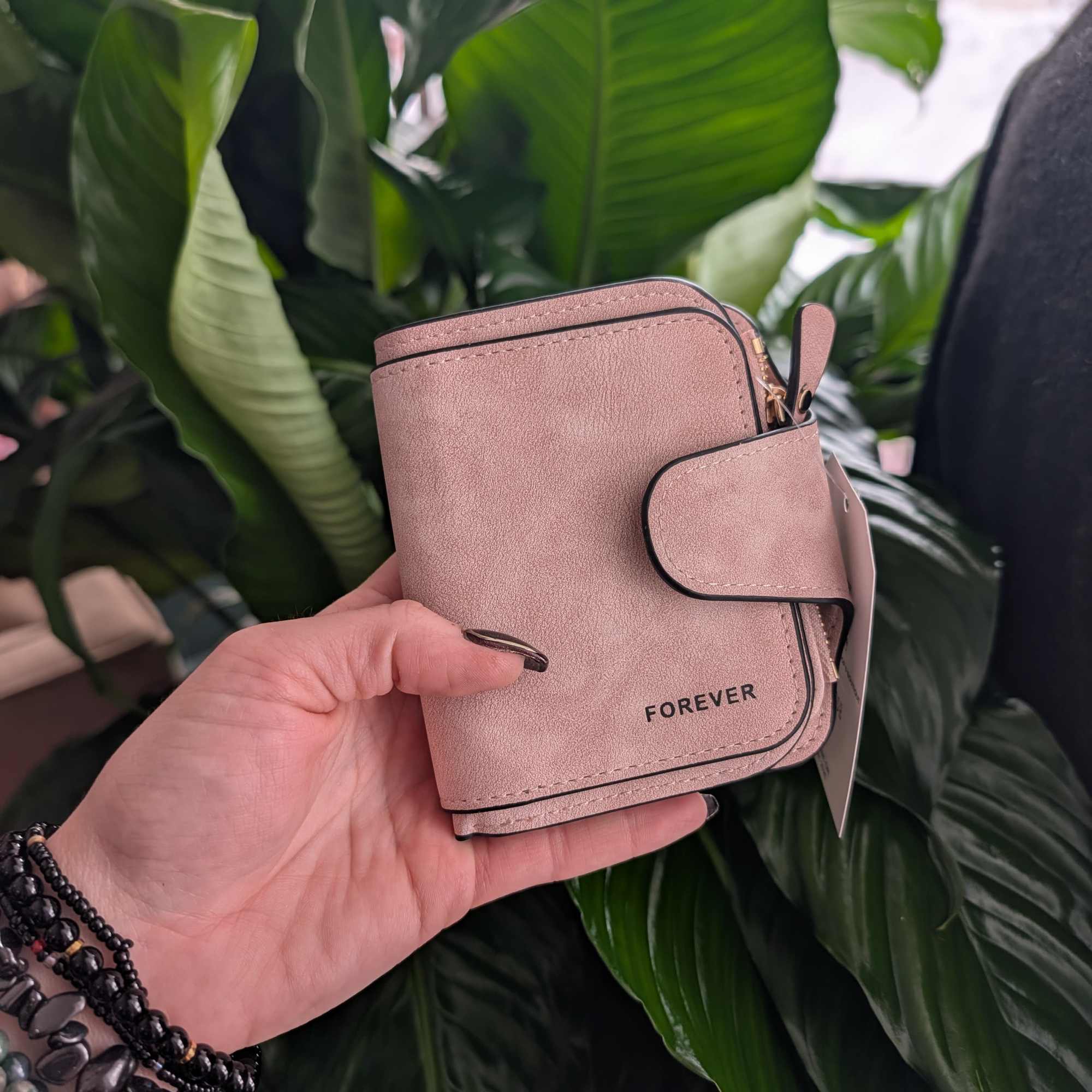 Hand holding a small pink wallet in front of large green leaves.