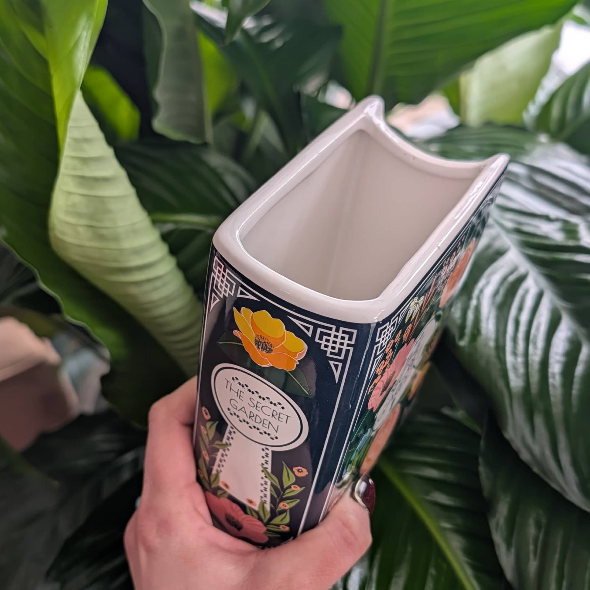 Hand holding a decorative ceramic vase with large green leaves behind it