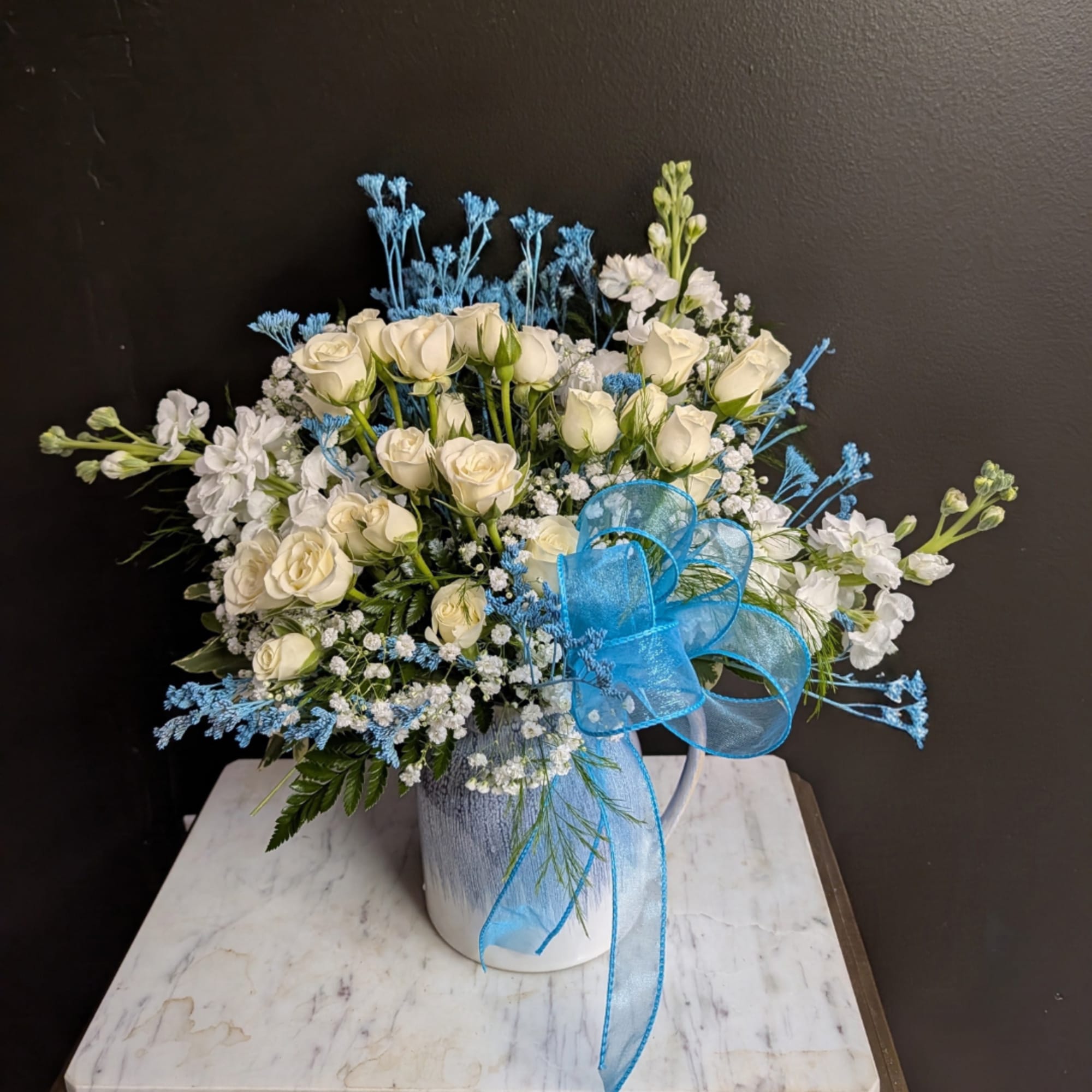 White roses and blue flowers in a white pitcher with a blue ribbon